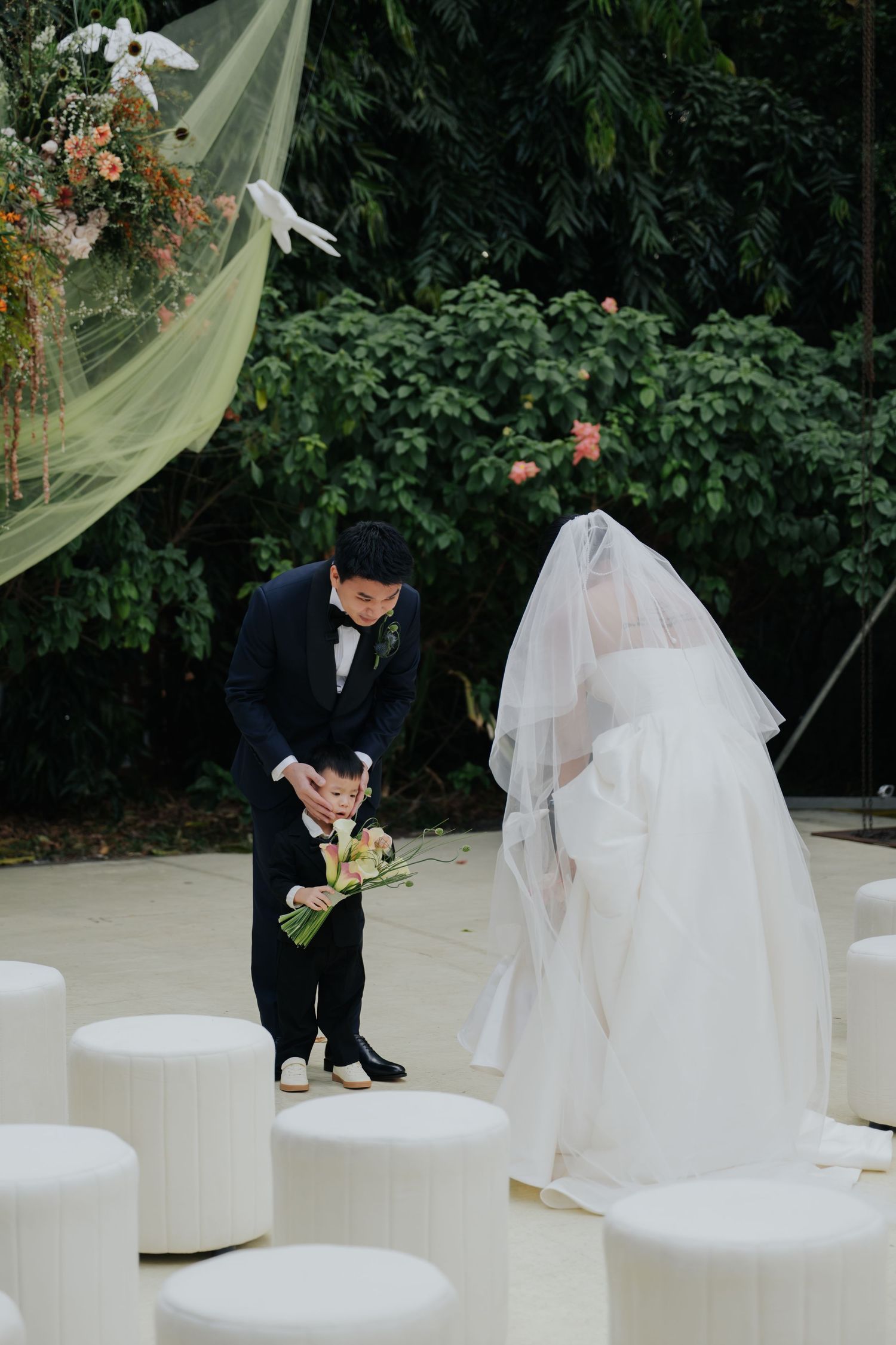 A bride and groom bow to each other during an outdoor wedding ceremony surrounded by lush greenery and white seating.
