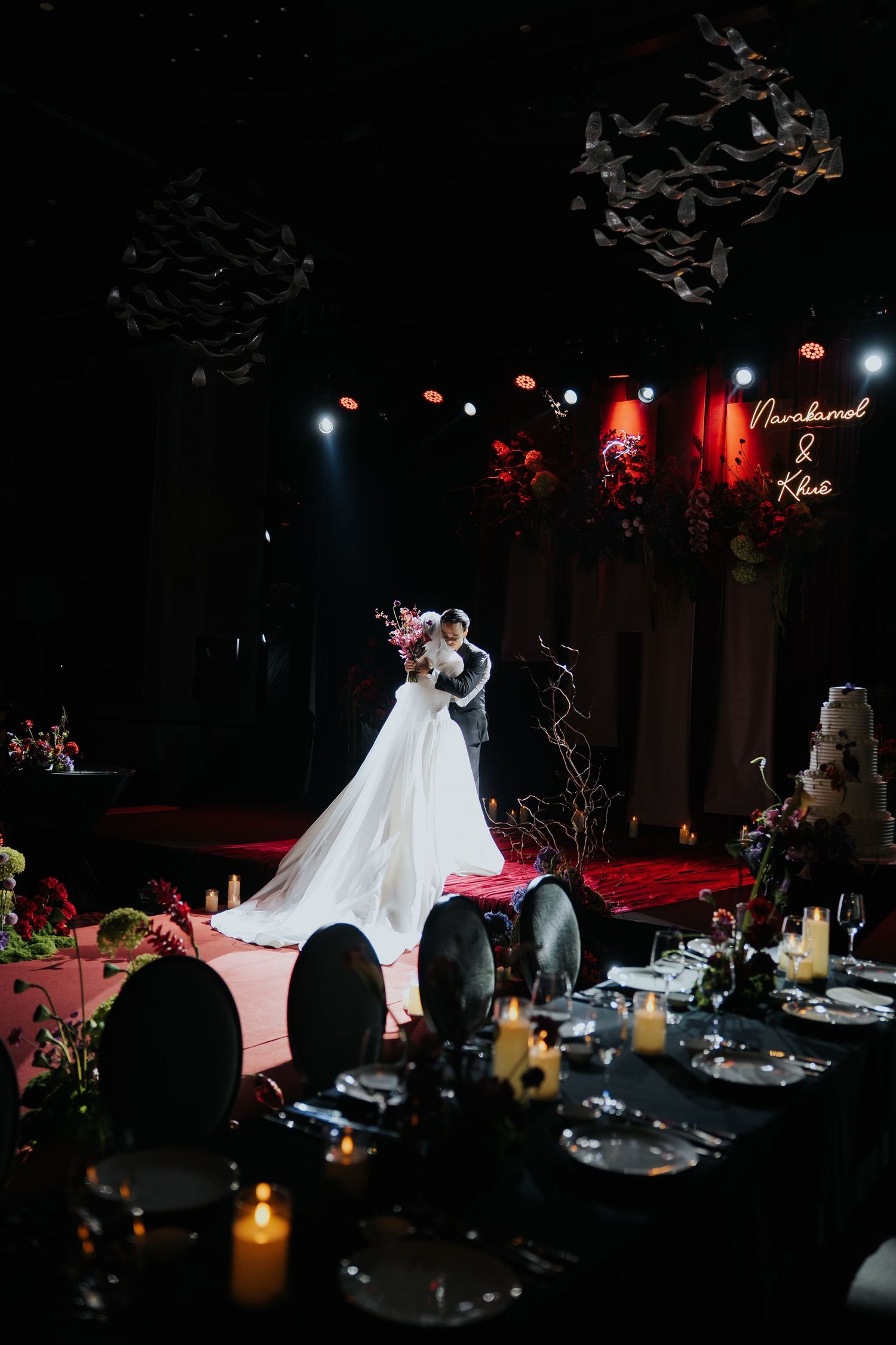 Bride and groom share first dance on illuminated stage at elegant dark-themed wedding reception with candlelit tables.