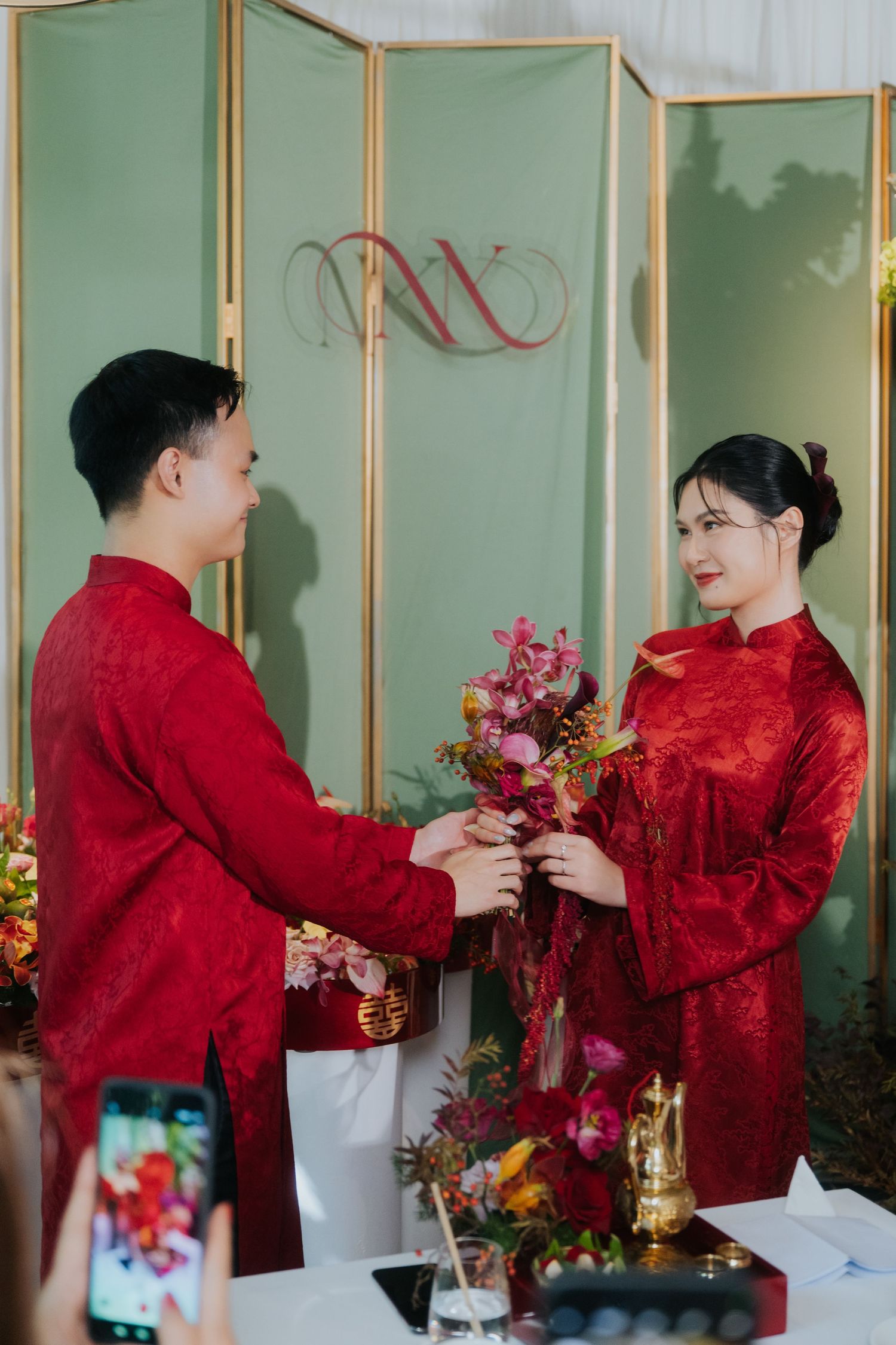 Couple in red traditional Chinese wedding attire exchanging flowers at a ceremony with a decorative screen backdrop.