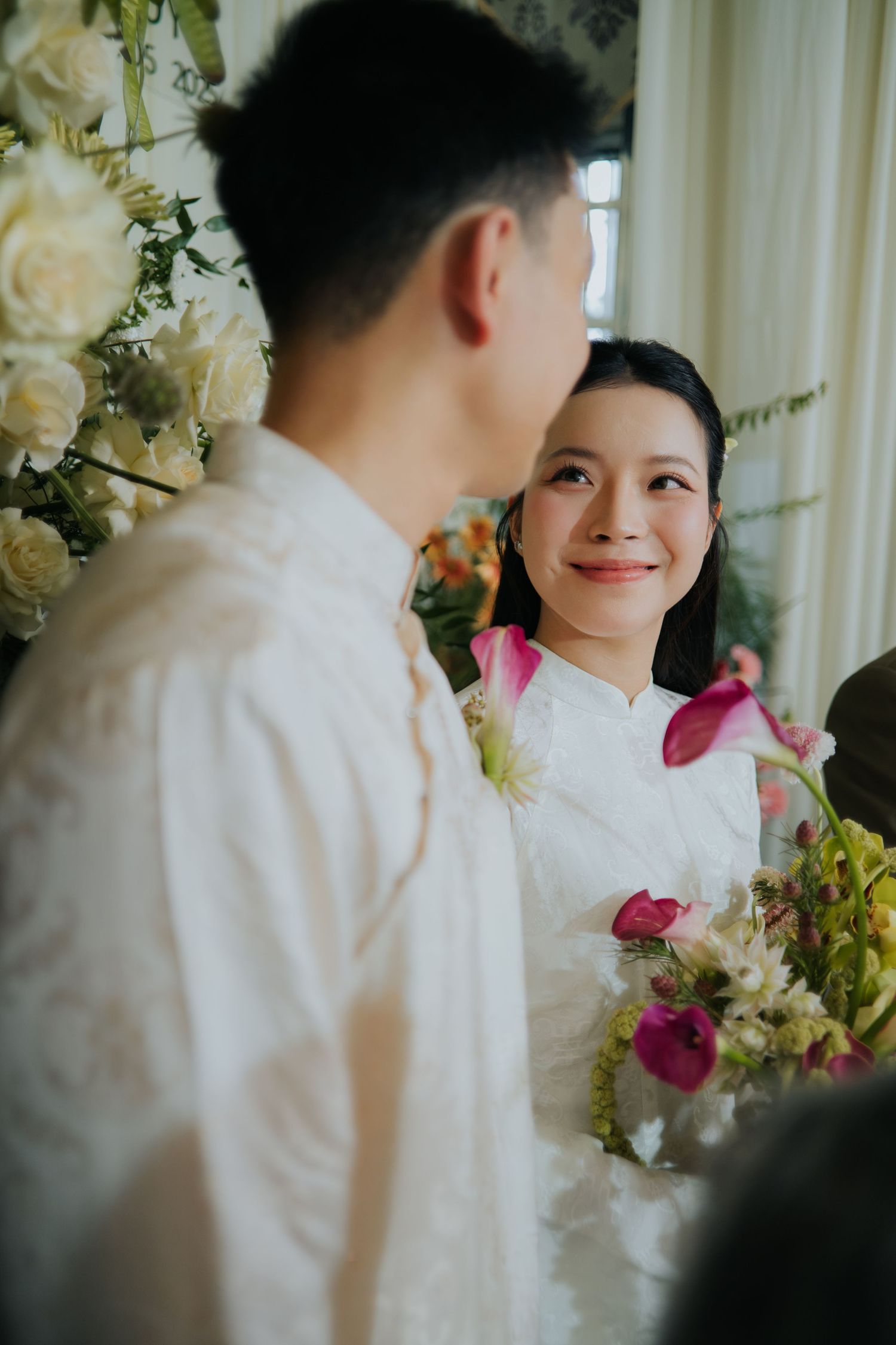A smiling bride in a white ao dai holds pink flowers while gazing at her groom during a traditional Vietnamese wedding ceremony.
