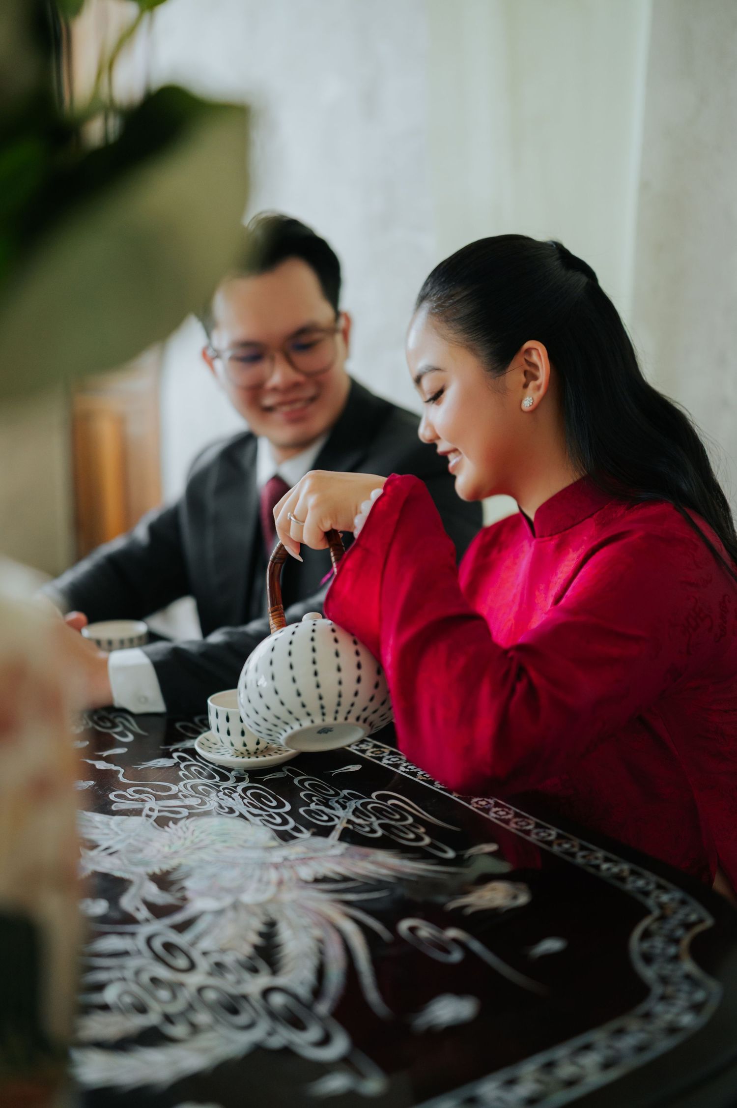 A woman in a red ao dai pours tea from a ceramic teapot at an ornate table during a traditional Vietnamese tea ceremony.