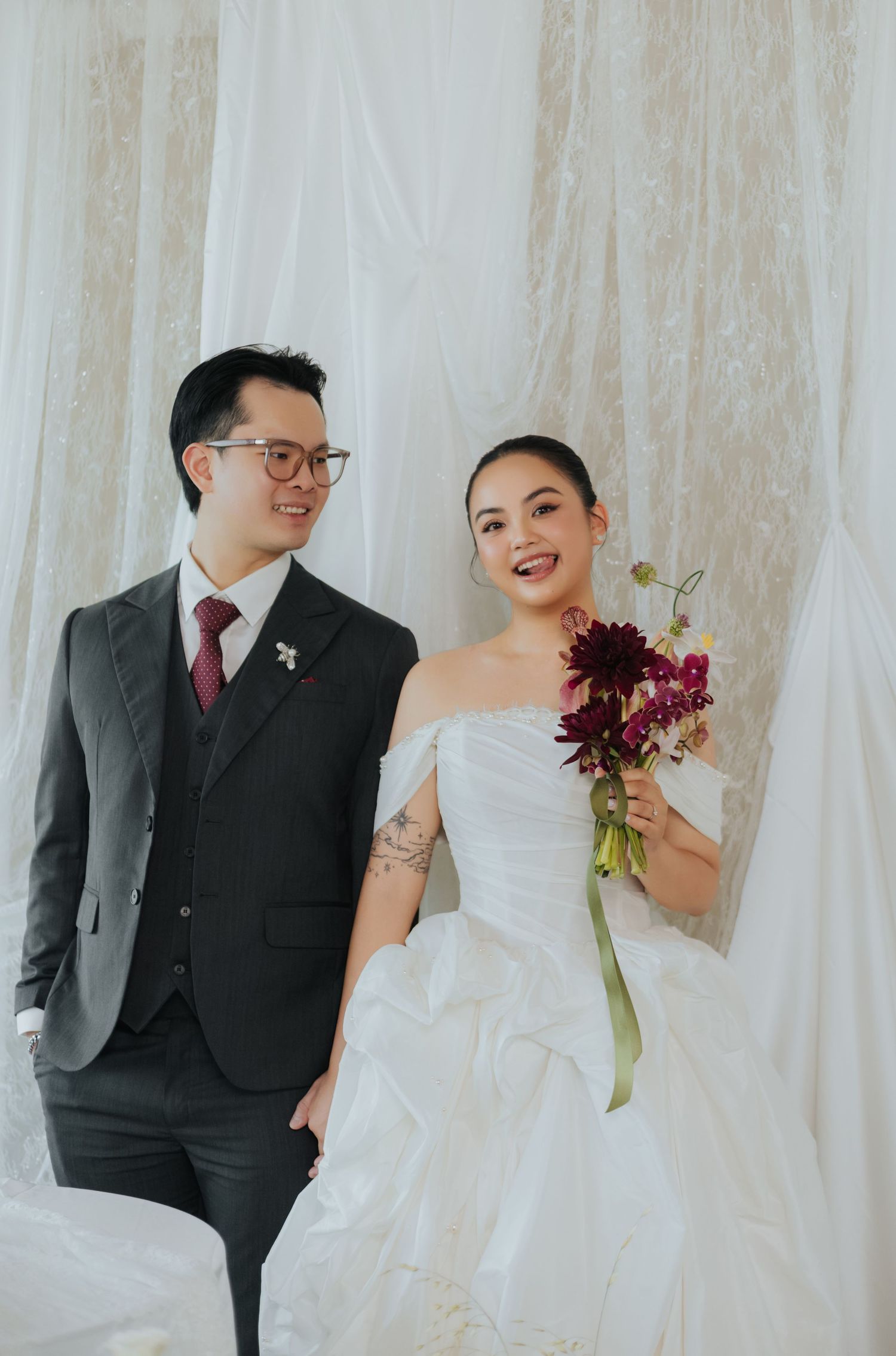 A smiling couple in wedding attire, groom in gray suit, bride in white gown holding burgundy flowers, posed before white drapes.