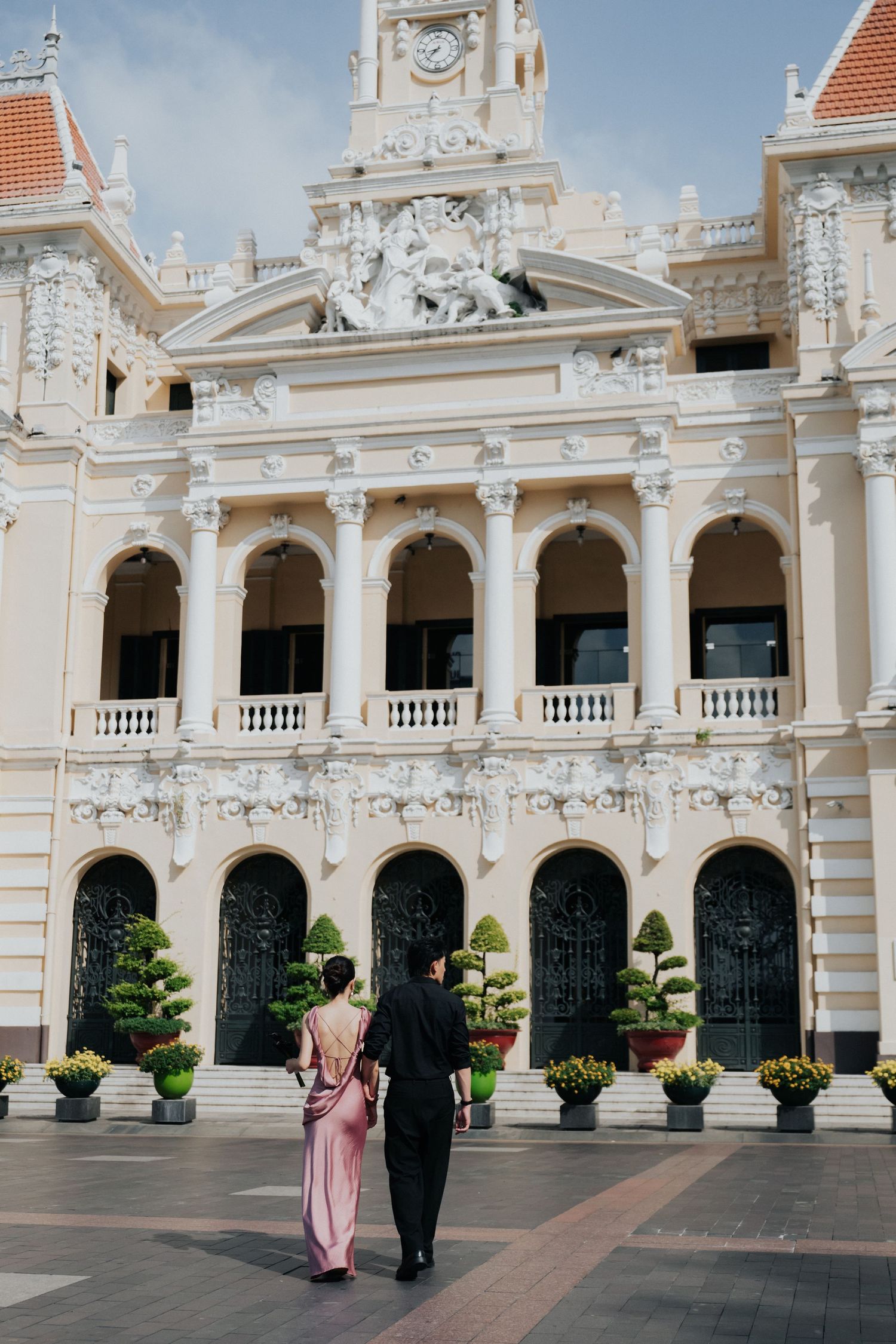 Couple in formal attire walking toward ornate French colonial building with arched facades and decorative sculptures in Ho Chi Minh City.