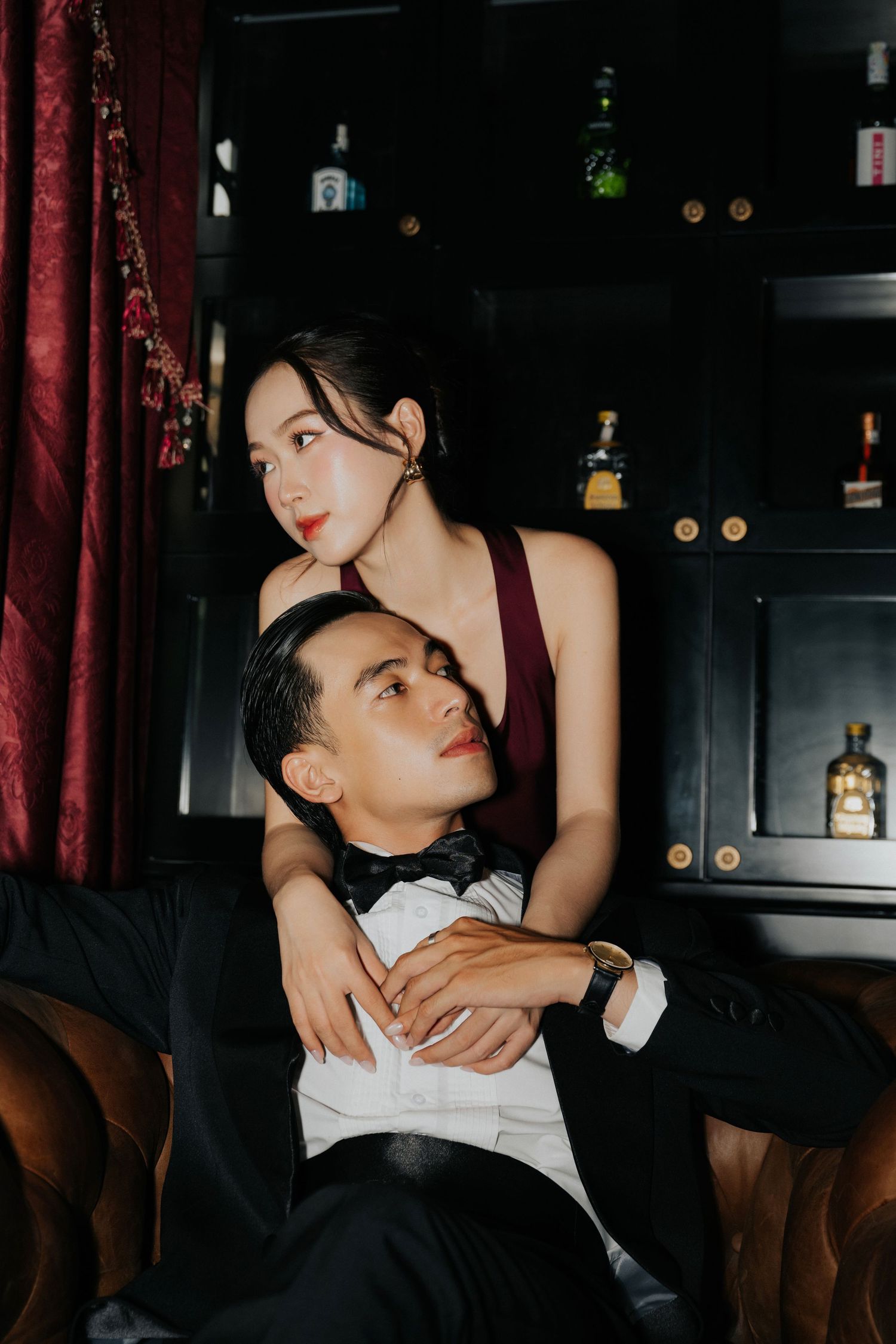 Elegant couple in formal attire pose intimately in a dark, sophisticated bar setting with liquor bottles in background.
