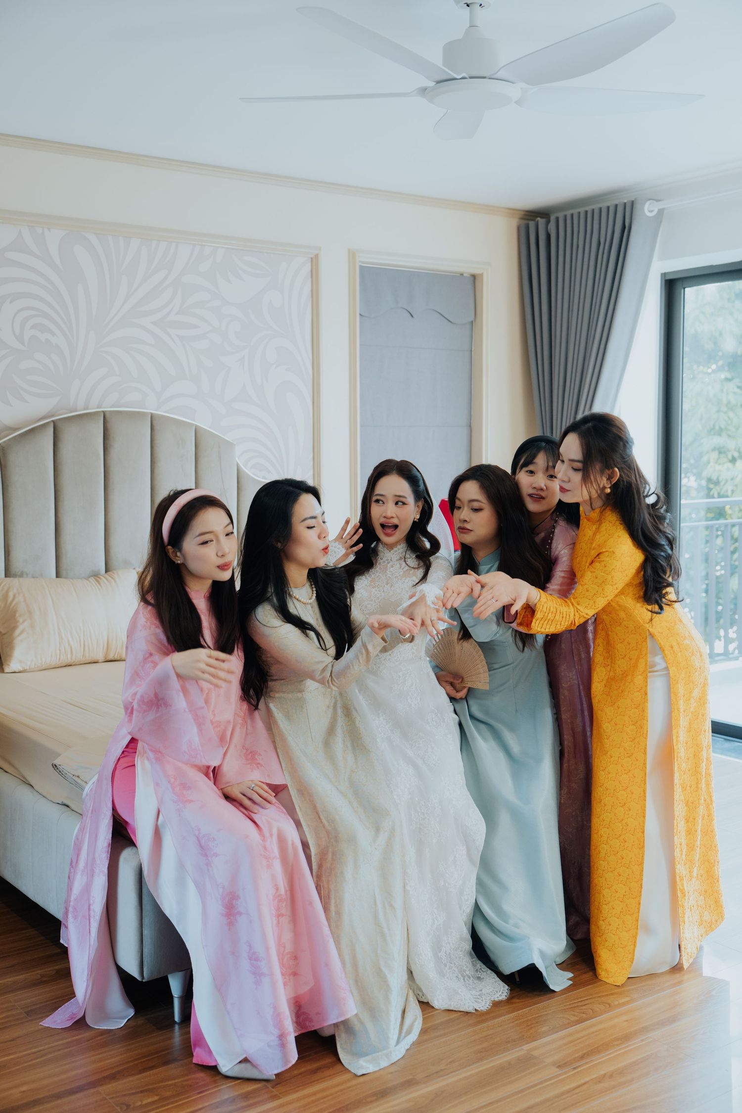 Six women in colorful Vietnamese ao dai dresses gather excitedly in a bright bedroom, celebrating with a bride in white.