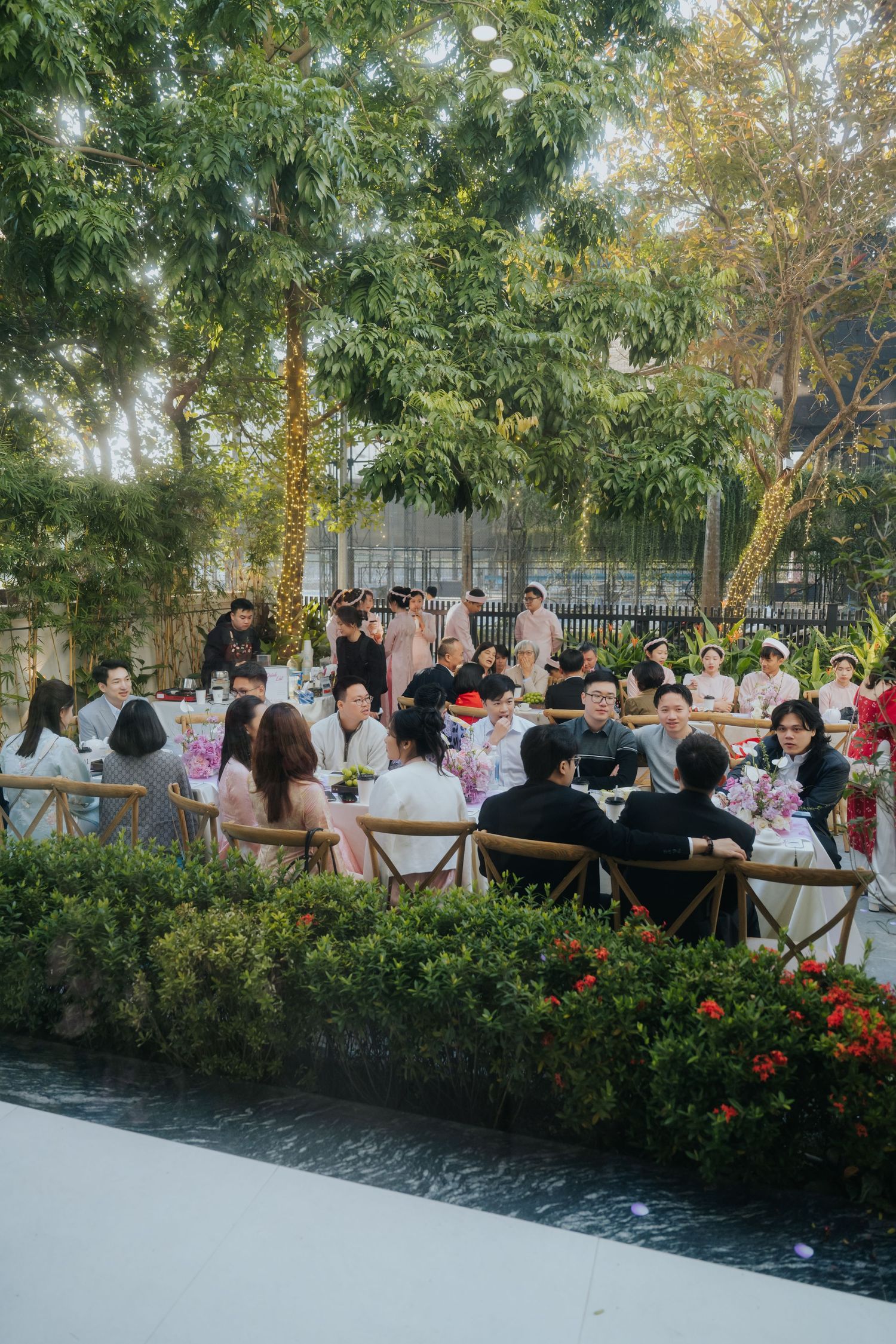 Guests gather at an elegant outdoor garden wedding reception, seated at decorated tables surrounded by lush green trees.