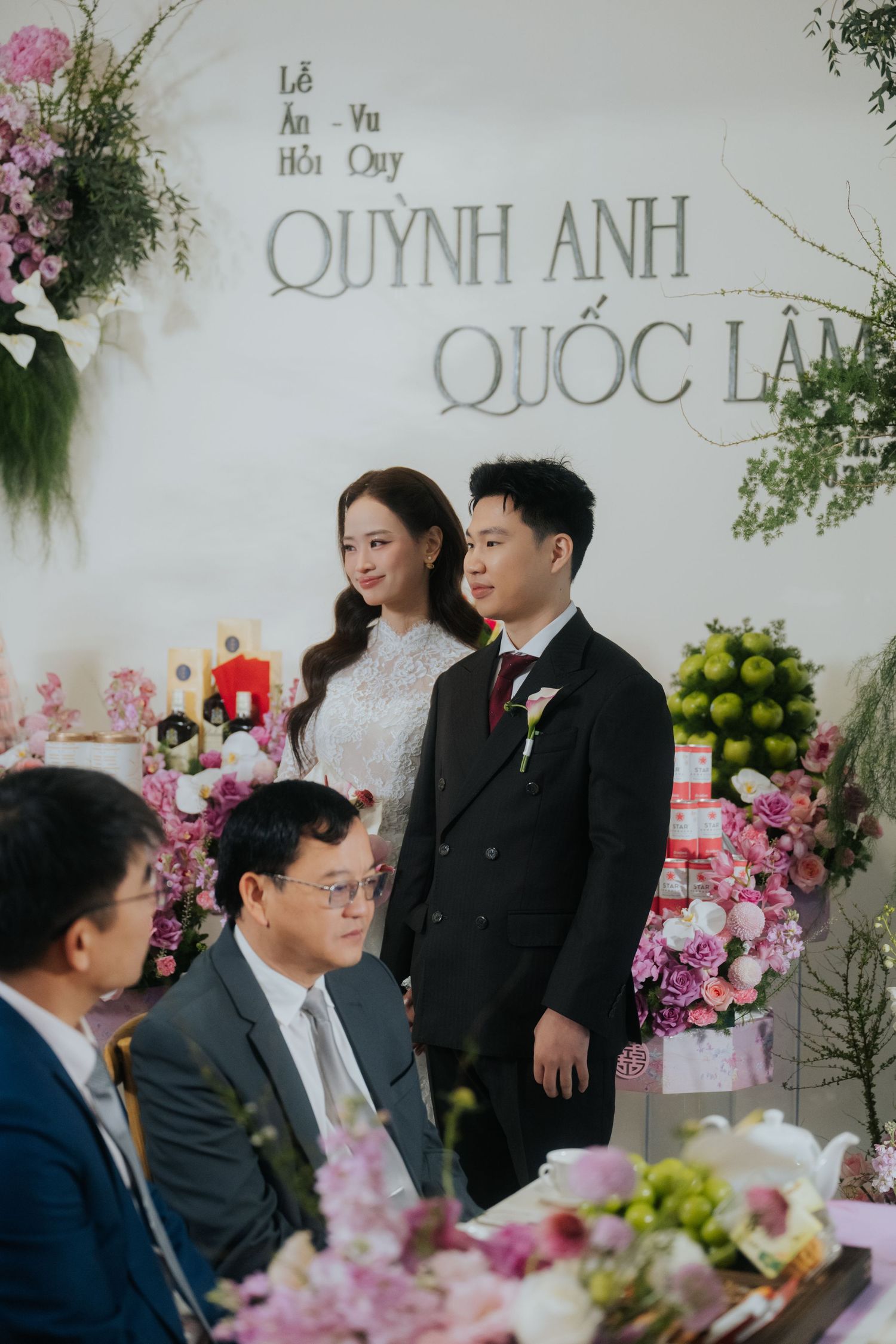 Vietnamese wedding ceremony with bride in white lace dress and groom in black suit standing before floral backdrop.