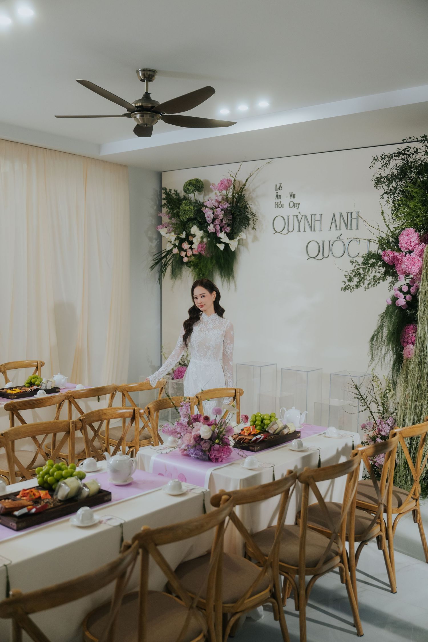 Bride in white lace dress stands at elegantly decorated wedding reception table with floral backdrop reading Quynh Anh.