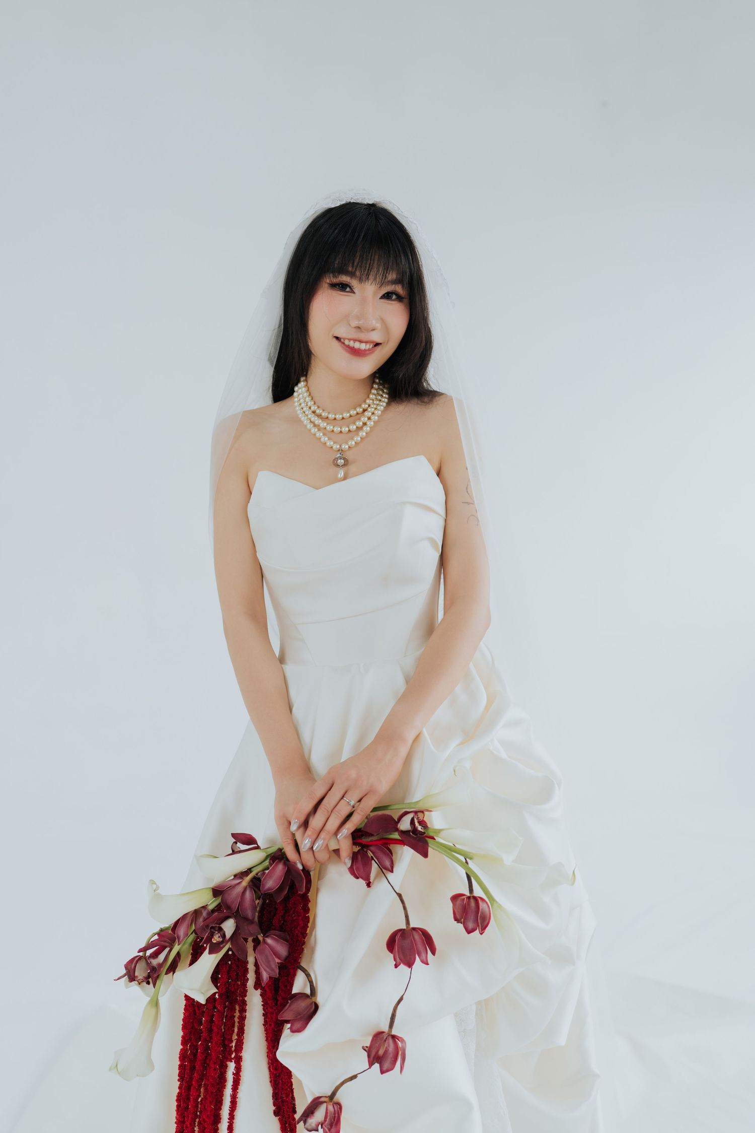 A smiling bride in a strapless white gown with pearl necklace holds dark red floral bouquet against white background.