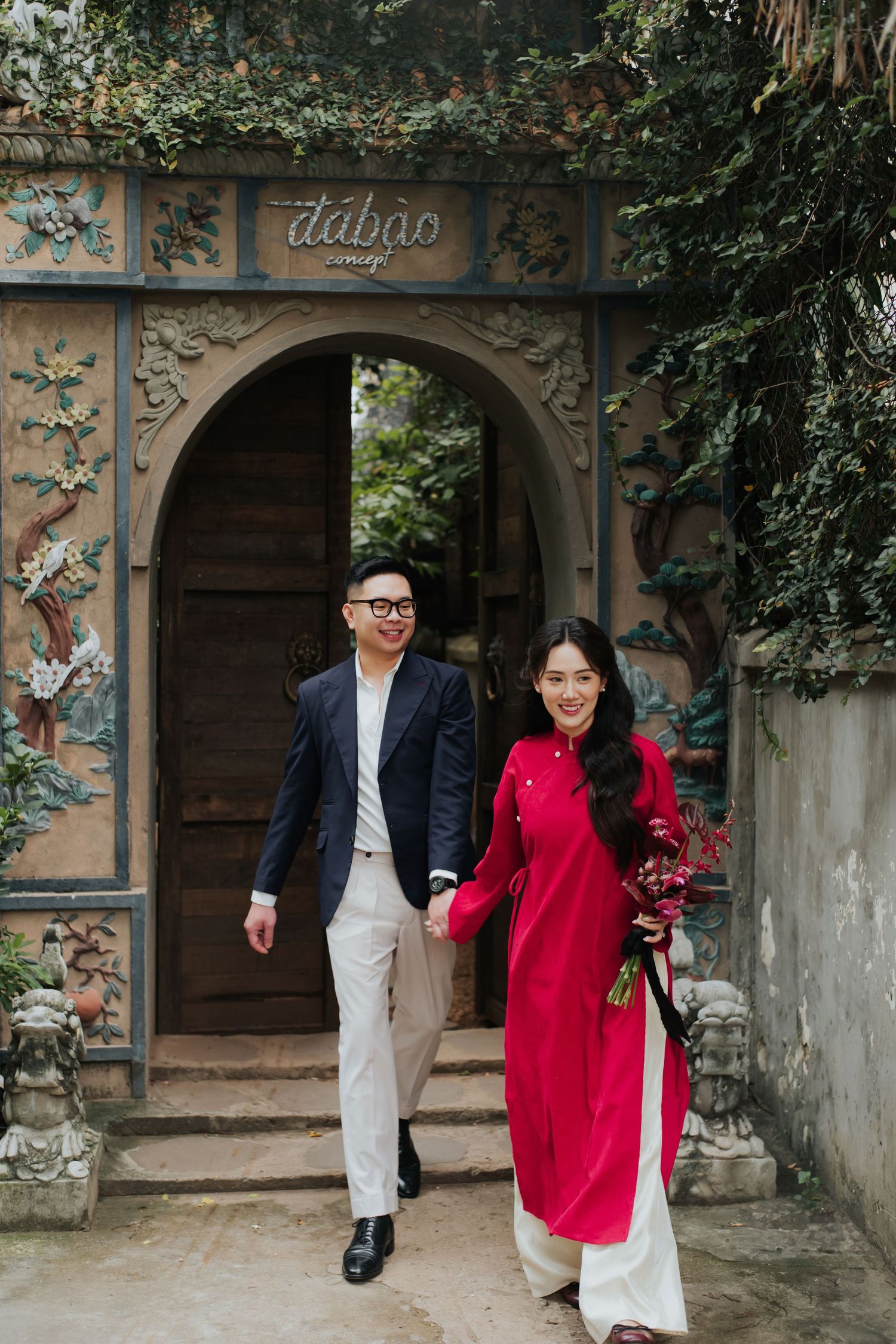 Couple in traditional Vietnamese ao dai and suit walking through ornate garden gate holding hands.