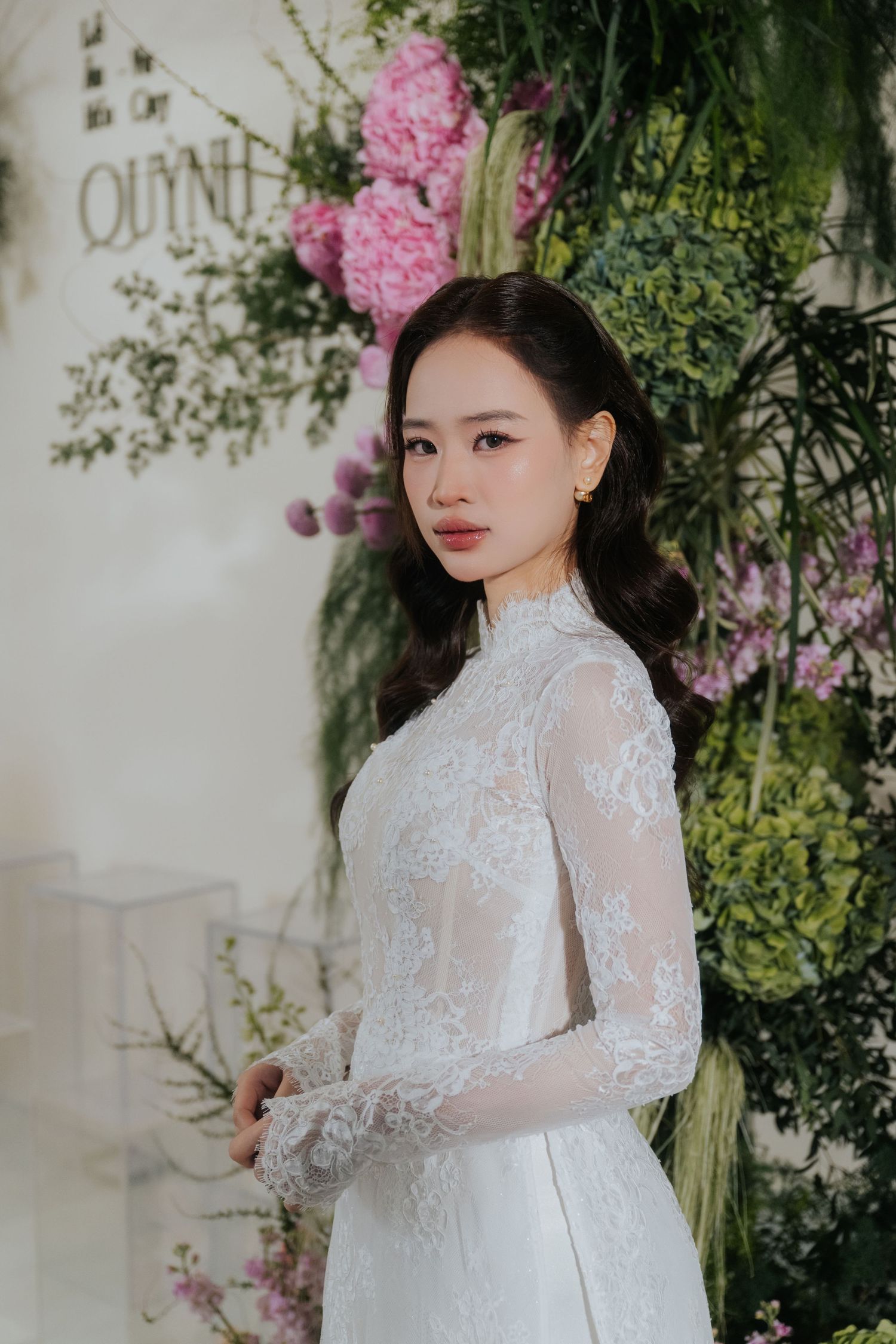 A young Asian woman in an elegant white lace high-neck bridal gown poses gracefully against a floral backdrop.