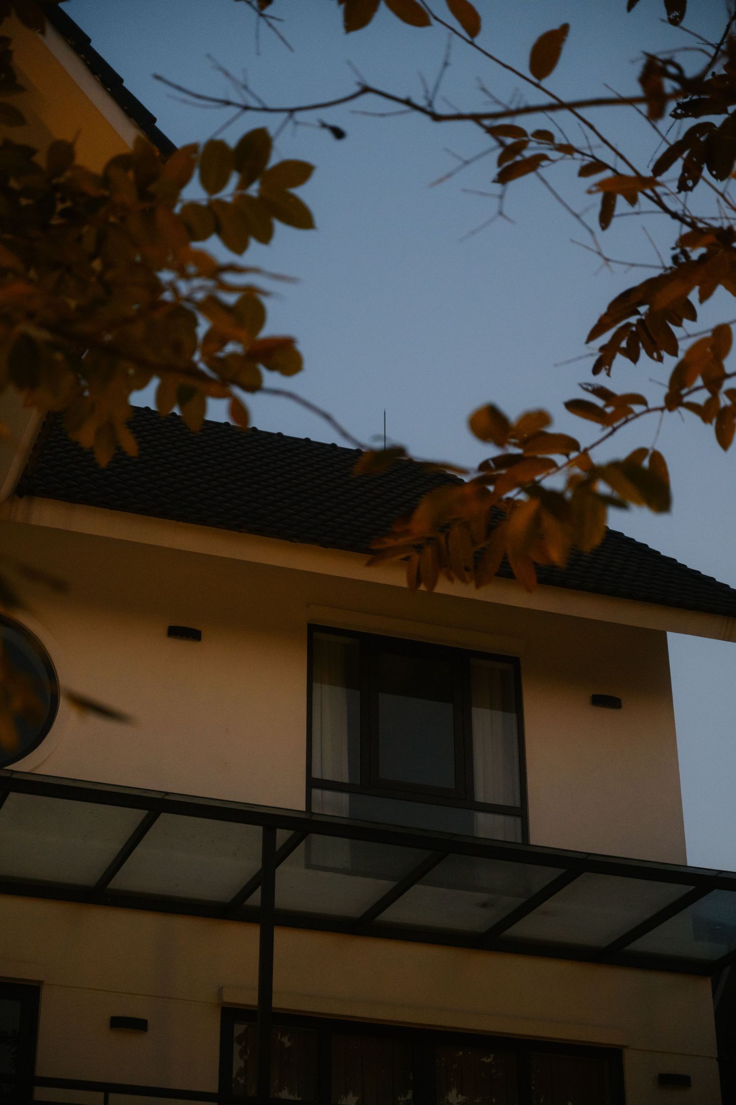 Autumn leaves frame a two-story house with dark roof tiles and a covered balcony under a clear dusk sky.