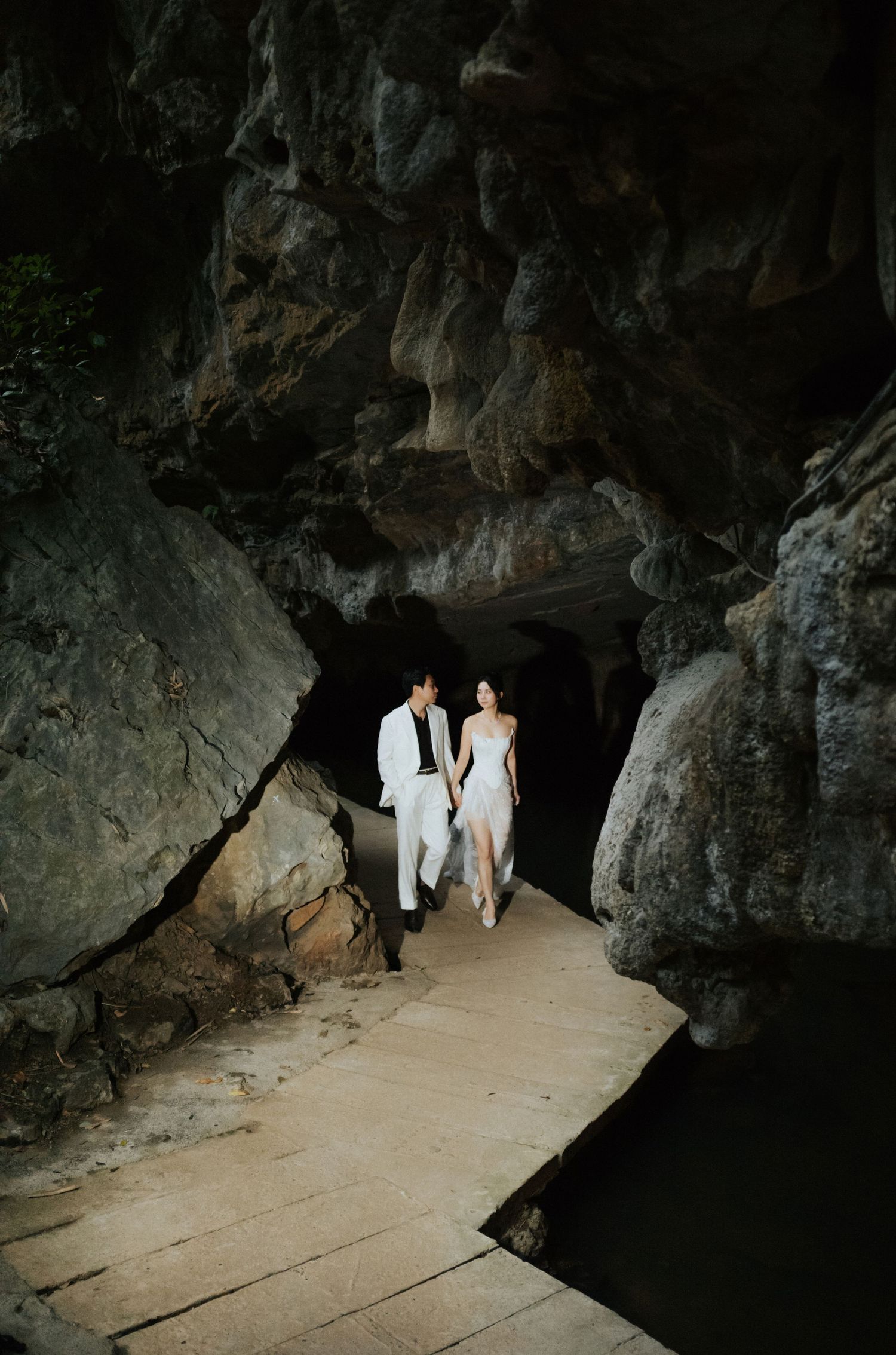 Couple in white attire walking through a dramatic cave passage with rocky walls and wooden pathway.