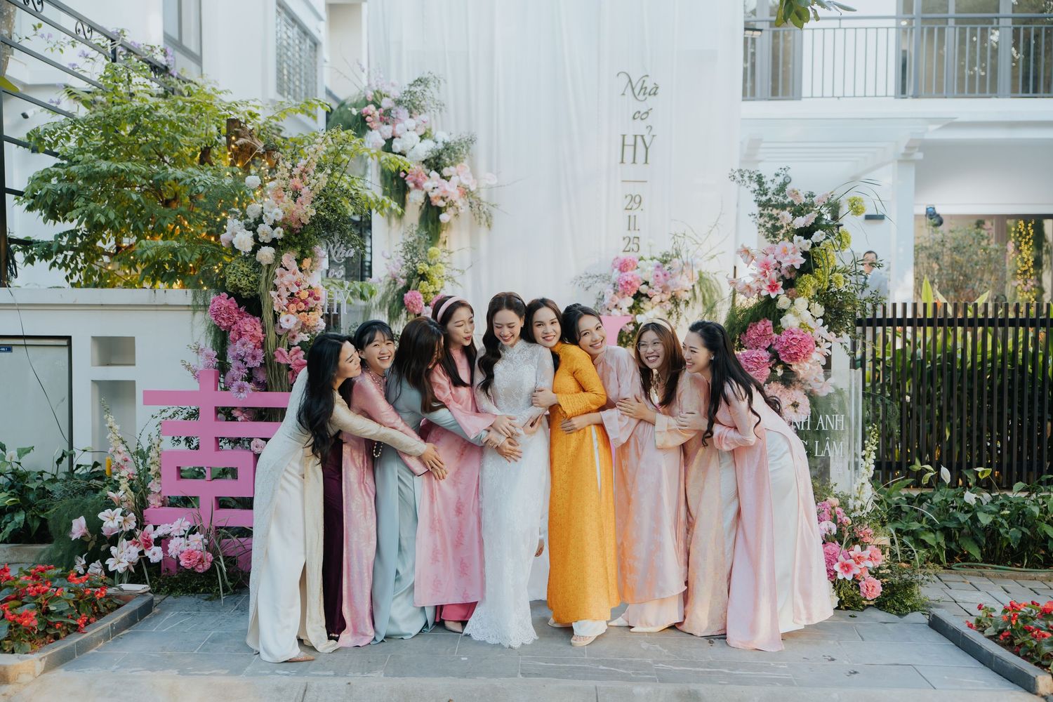 A bride in yellow ao dai poses with bridesmaids in pastel dresses at a floral Vietnamese wedding backdrop.