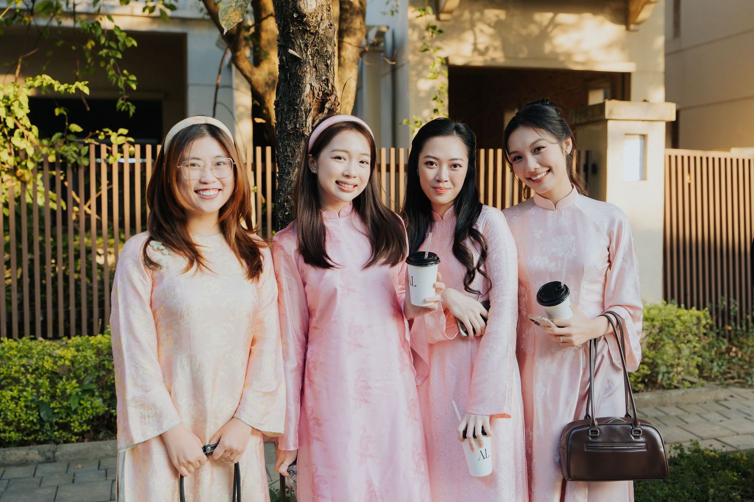 Four young women in matching pink ao dai traditional Vietnamese dresses smiling together outdoors.