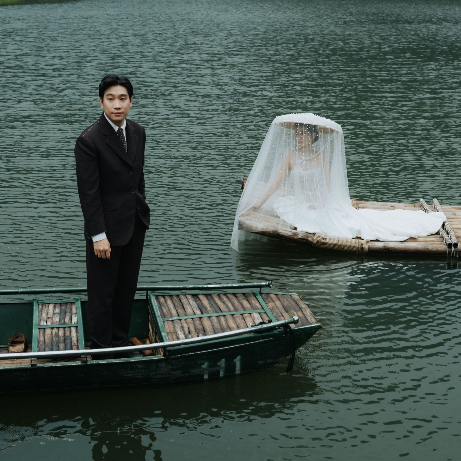 Man in black suit standing on a wooden boat on a calm green lake with a white draped object floating nearby.