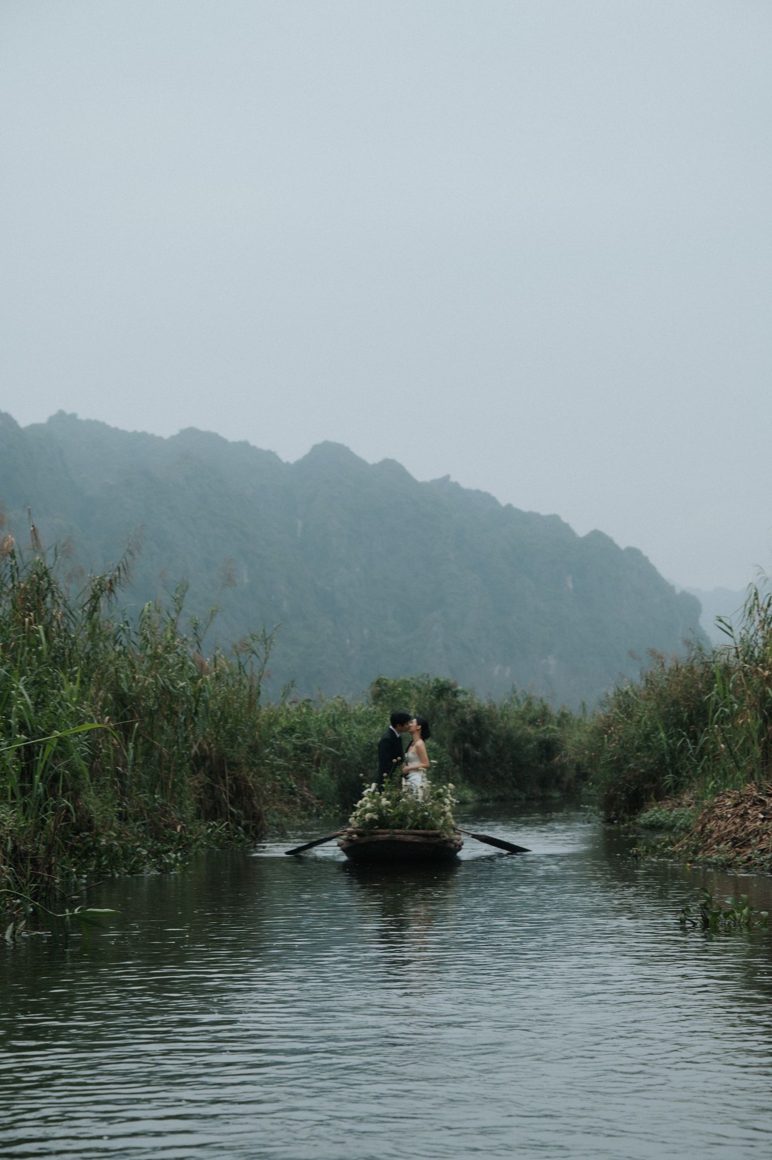 Couple standing in a flower-adorned wooden boat on a misty river surrounded by lush greenery and karst mountains.