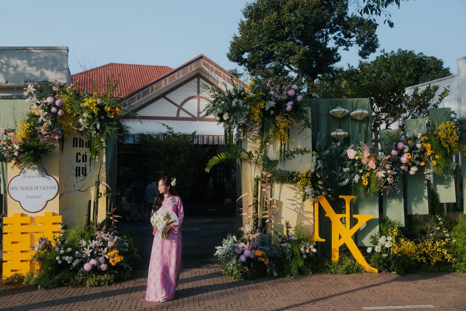 Woman in purple ao dai stands before a floral-decorated wedding venue entrance with yellow and green arch decor.