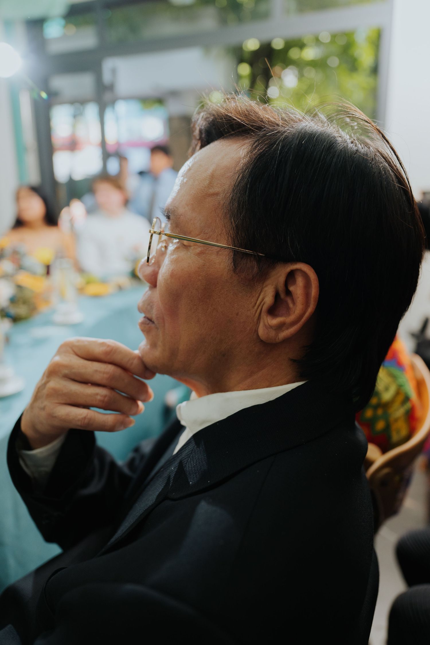 An elderly Asian man in a black suit and glasses sits thoughtfully at a formal dining event, hand raised to his chin.