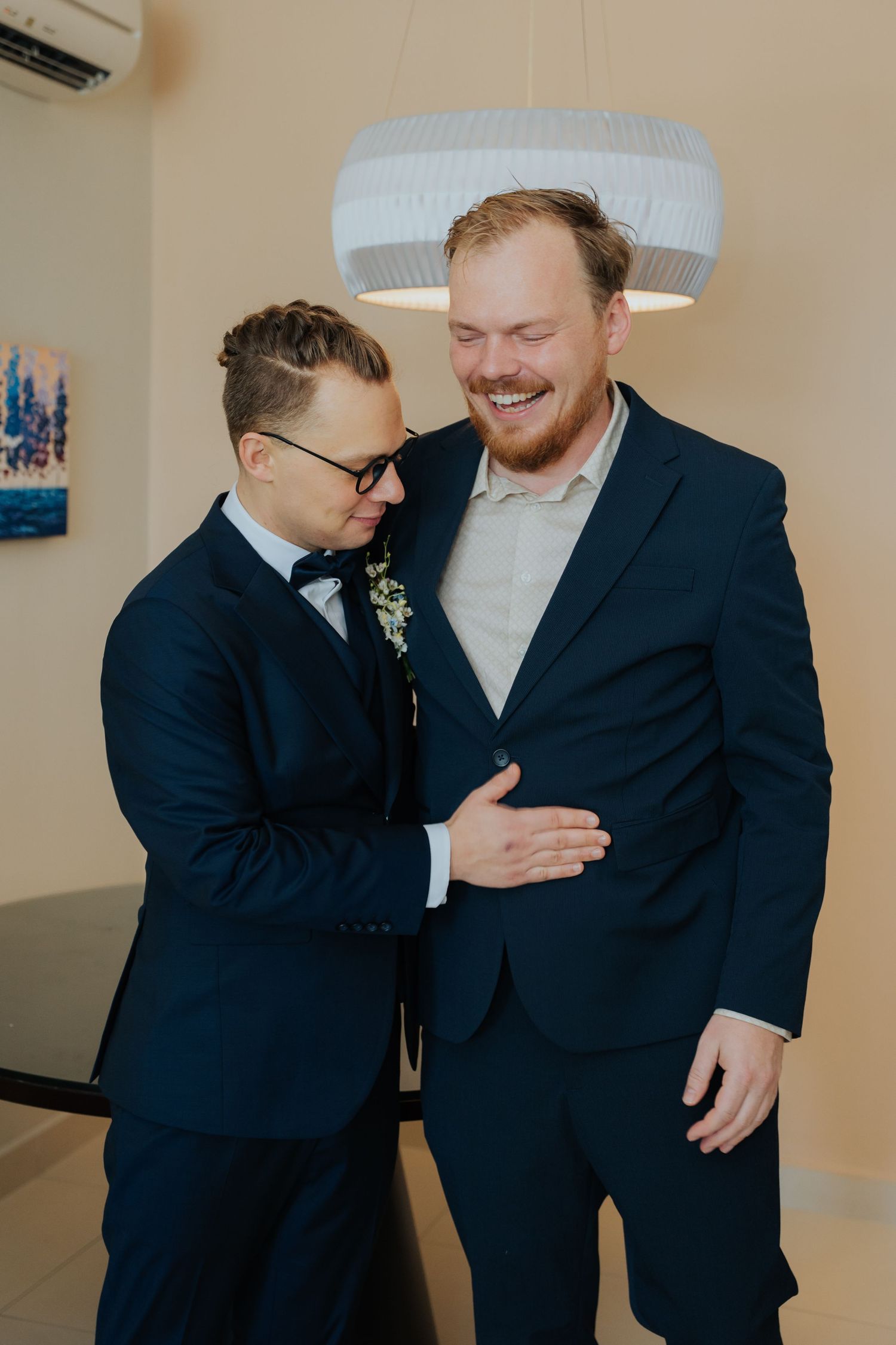 Two grooms in navy suits share a joyful moment, one smiling broadly as the other adjusts his jacket.