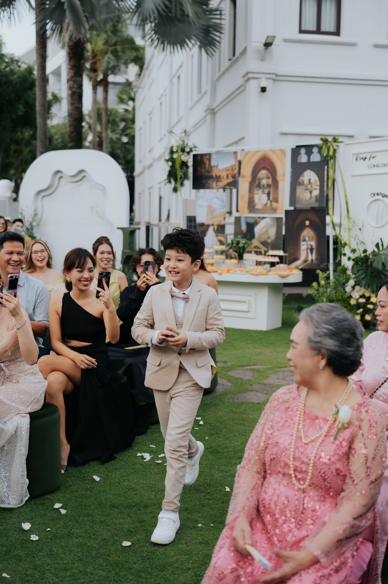 A young boy in a beige suit walks down the aisle at an outdoor wedding ceremony, smiling at guests.
