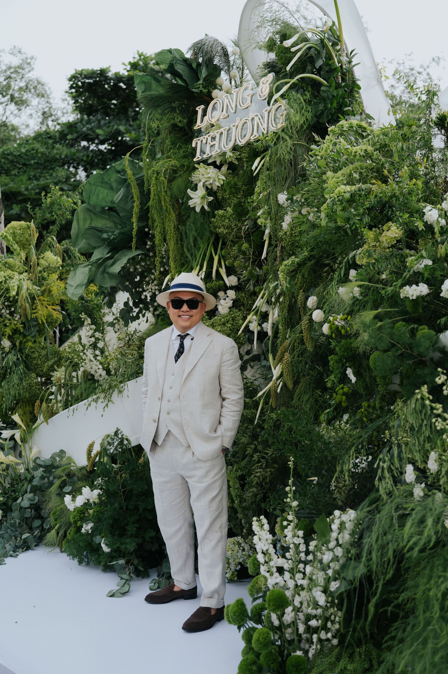 Man in beige suit and hat poses before lush green floral wedding backdrop with white flowers and Long Thuong sign.