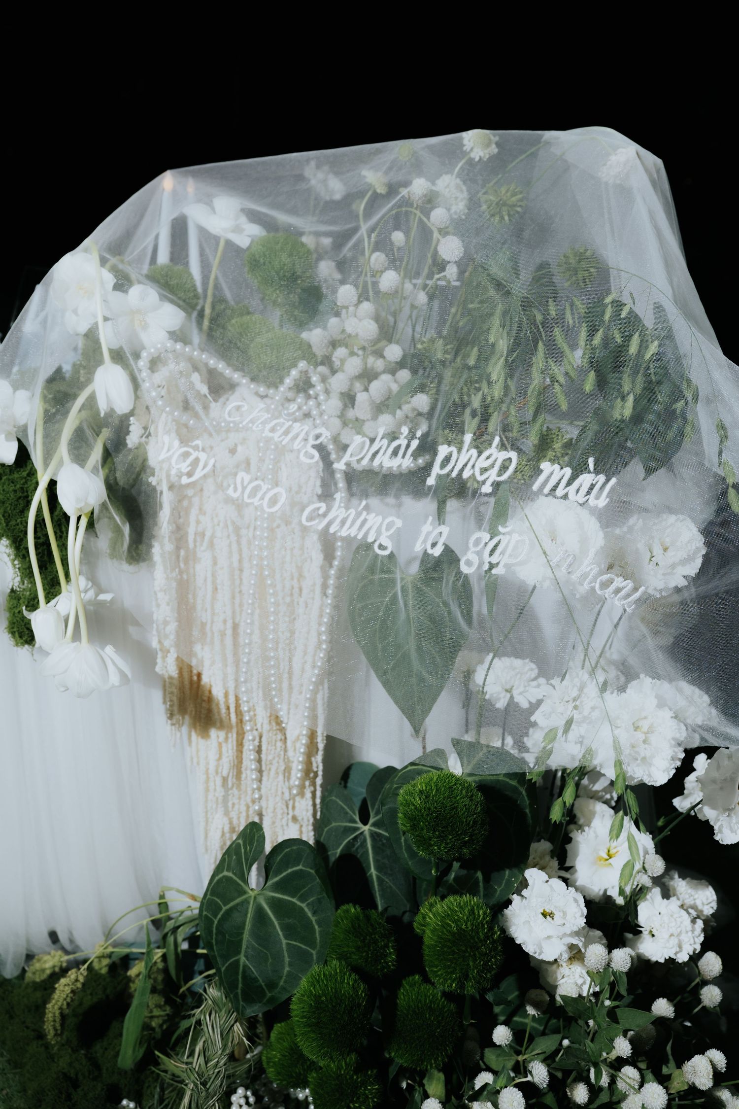 White floral wedding backdrop with sheer veil, pearl strands, and Vietnamese script on dark background.