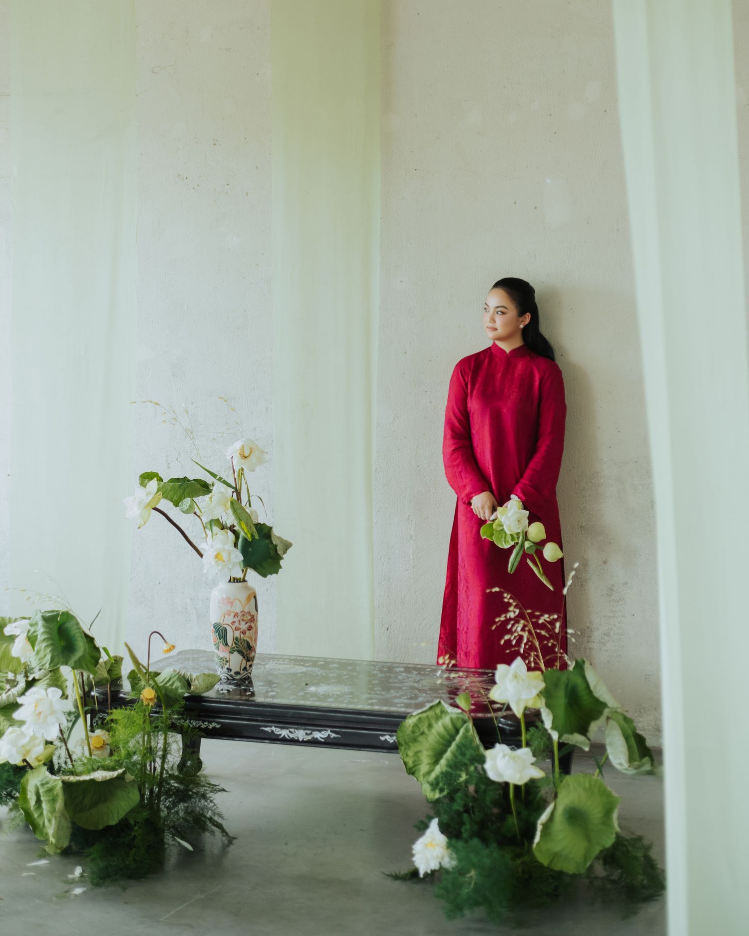 Woman in red ao dai dress holds white lotus flowers beside a decorated table with floral arrangements against a pale wall.