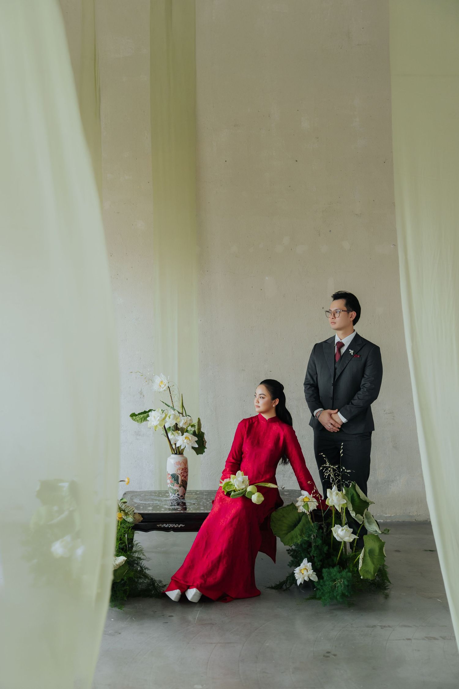 A woman in a red ao dai sits beside a man in a dark suit, surrounded by white flowers in an elegant indoor setting.