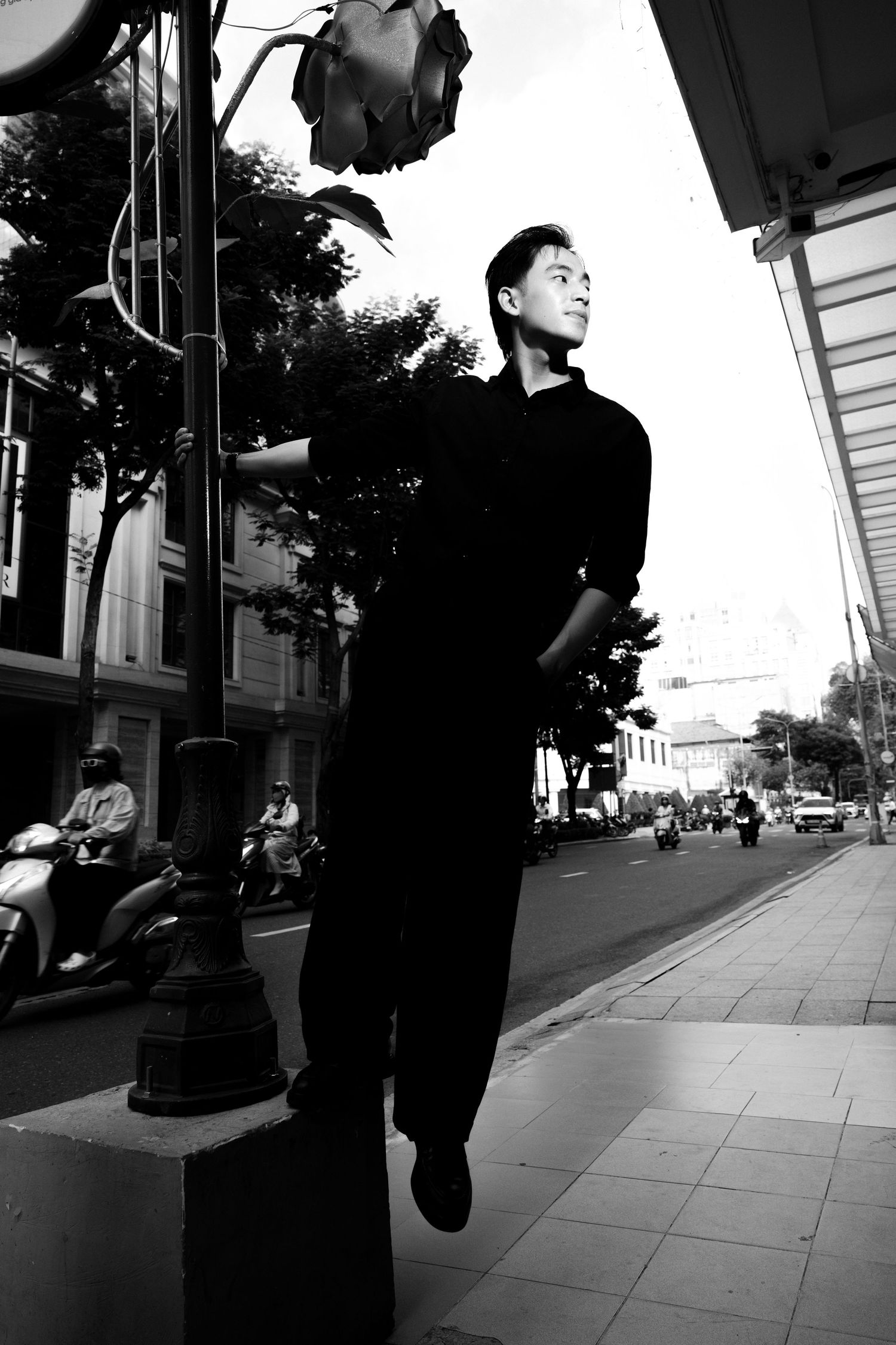 Black and white photo of a young Asian man in all black leaning against a street pole on a busy urban sidewalk.