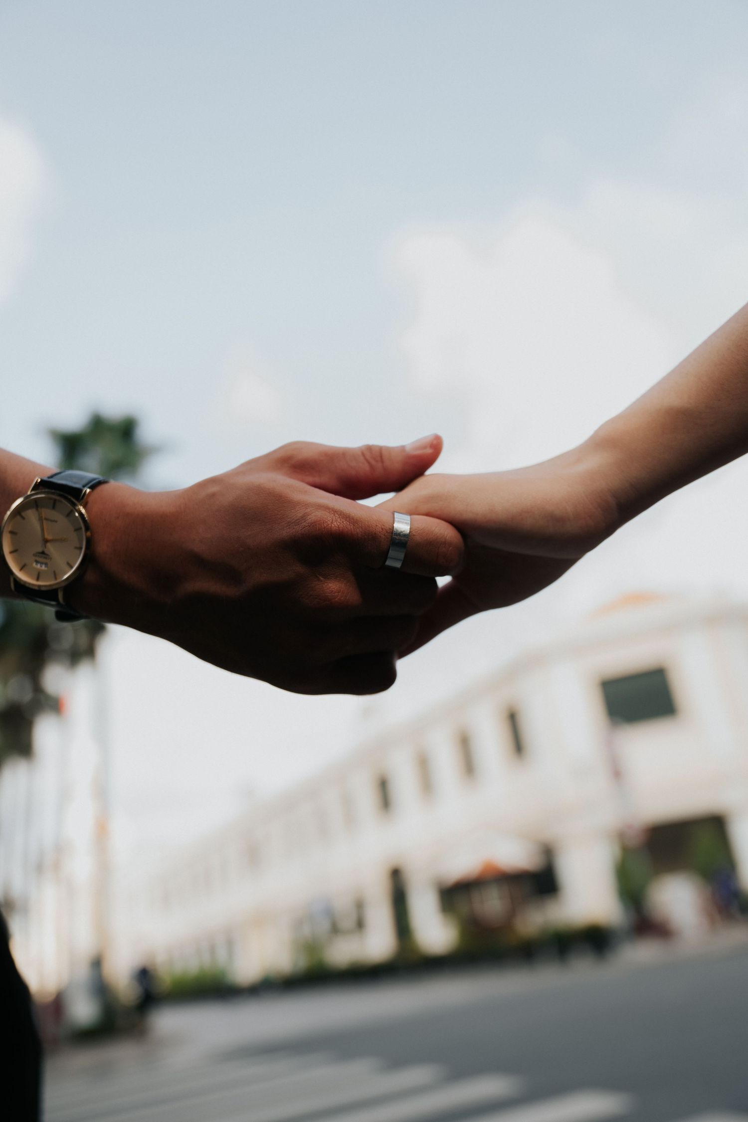 Close-up of two hands almost touching, one wearing a watch and the other a silver ring, with a blurred street background.