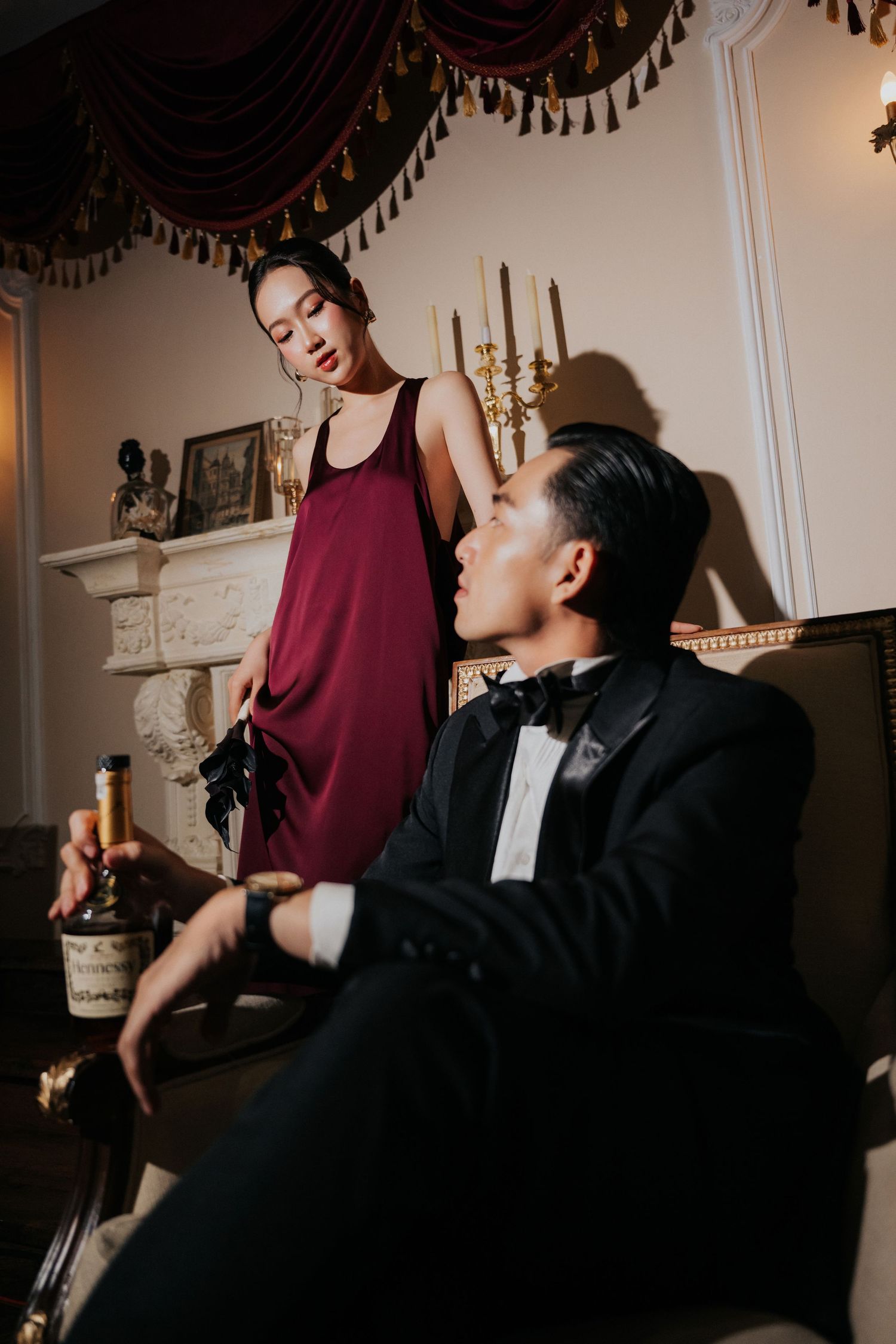Elegant couple in luxury room, woman in burgundy dress stands by fireplace, man in tuxedo holds champagne bottle.