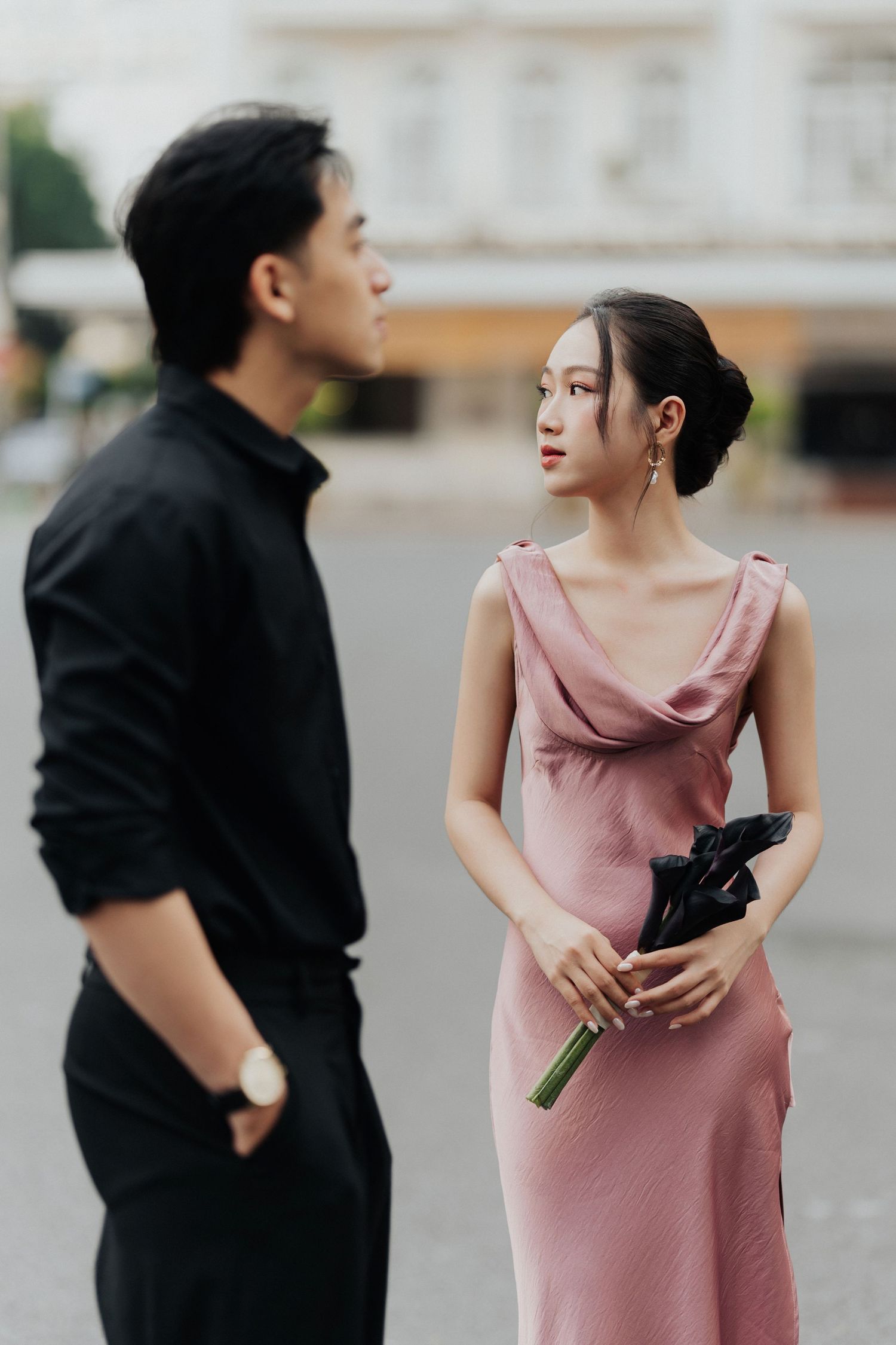 A young Asian couple in elegant attire, woman in pink dress holding flowers, sharing a moment outdoors.