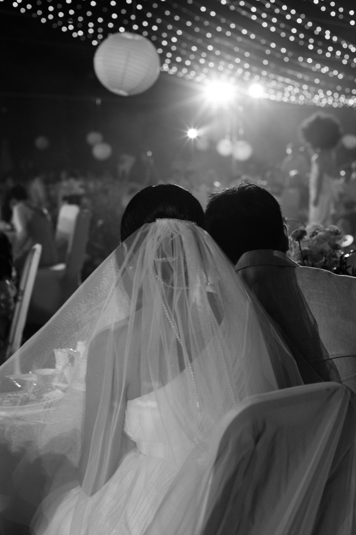 Black and white photo of bride and groom leaning together under wedding veil at elegant reception.