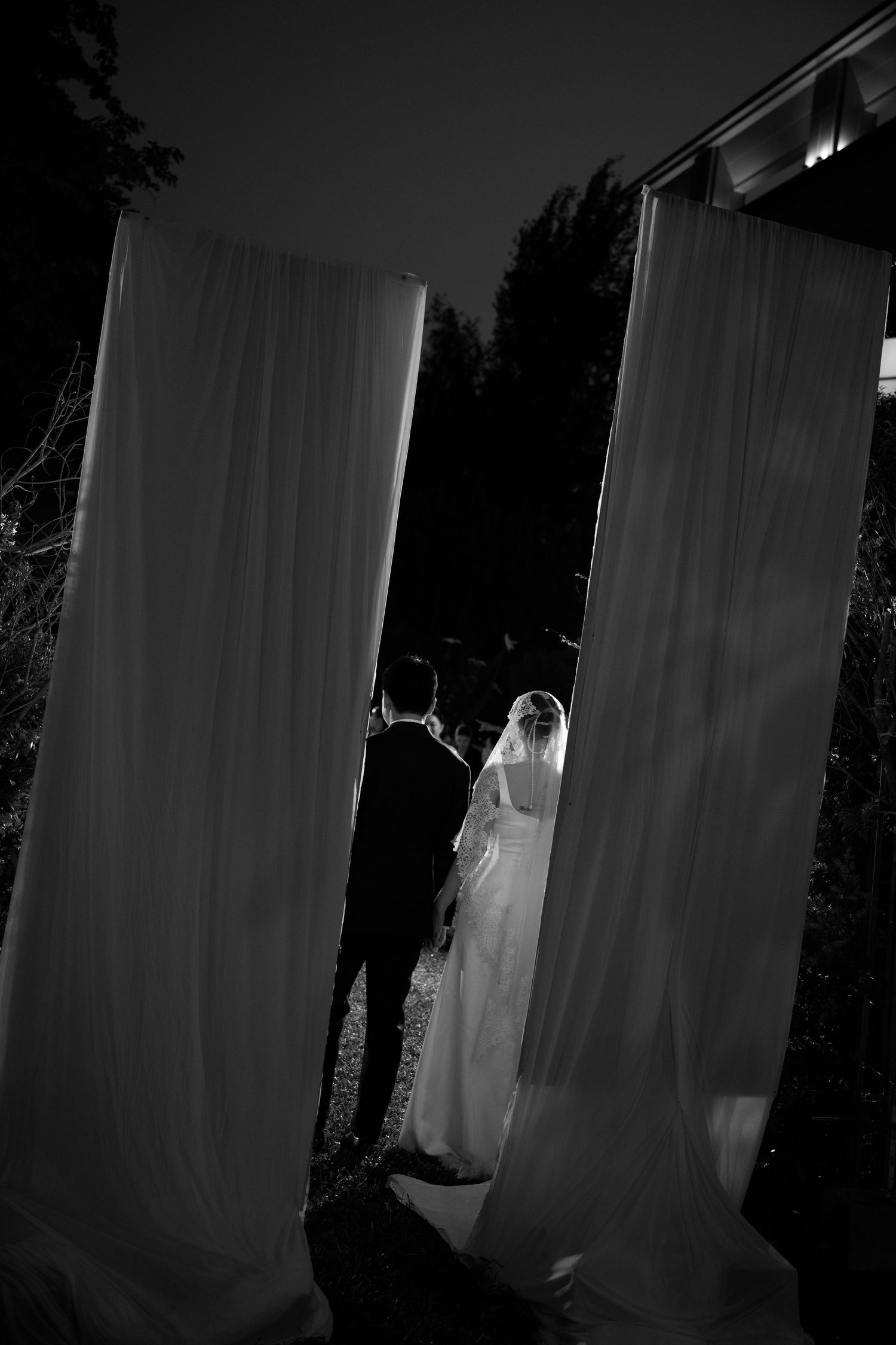 Black and white photo of bride and groom walking between white draped curtains at an outdoor evening wedding ceremony.