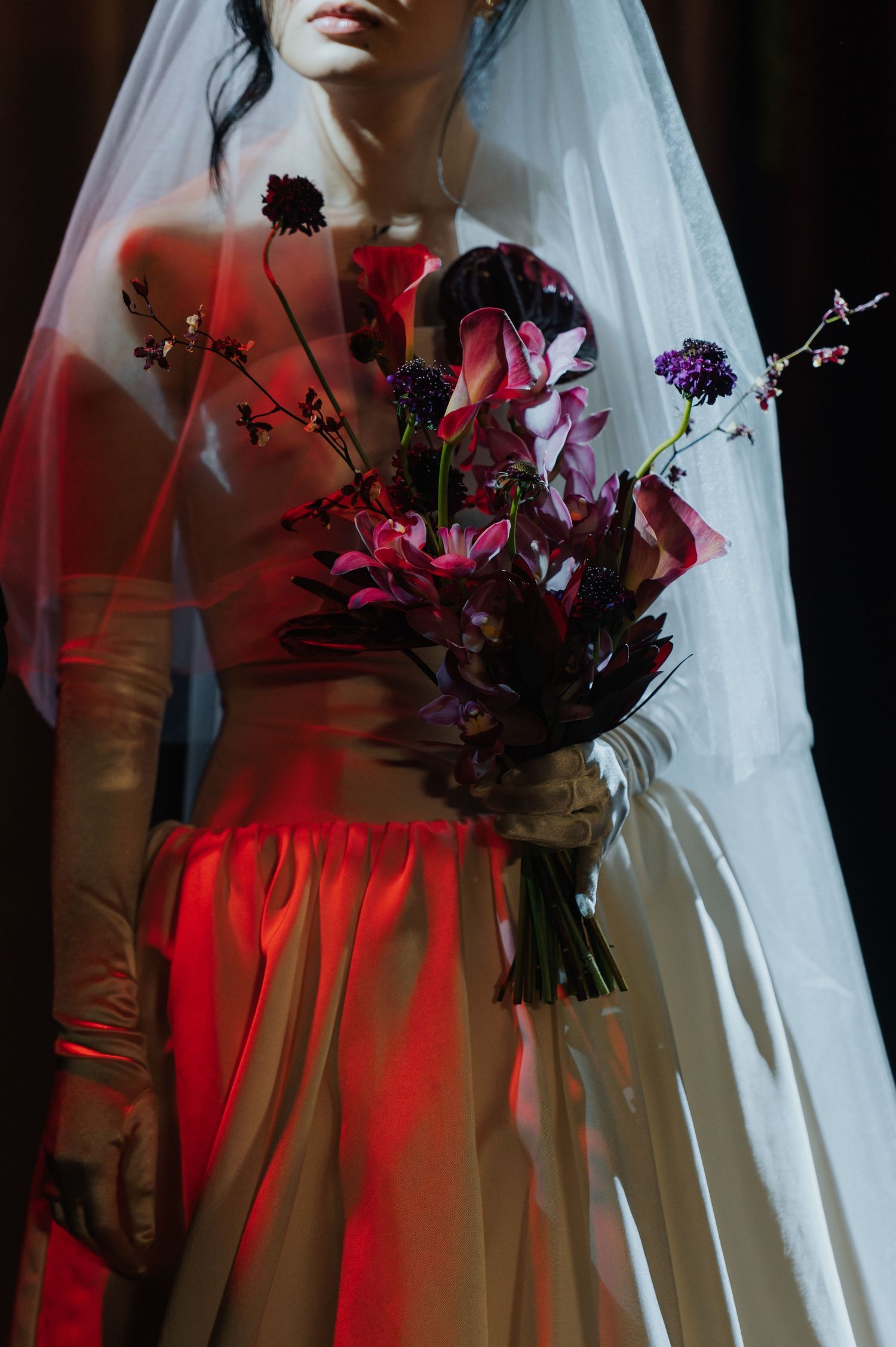 Bride in white gown and veil holds dark floral bouquet with pink and purple blooms under dramatic red lighting.
