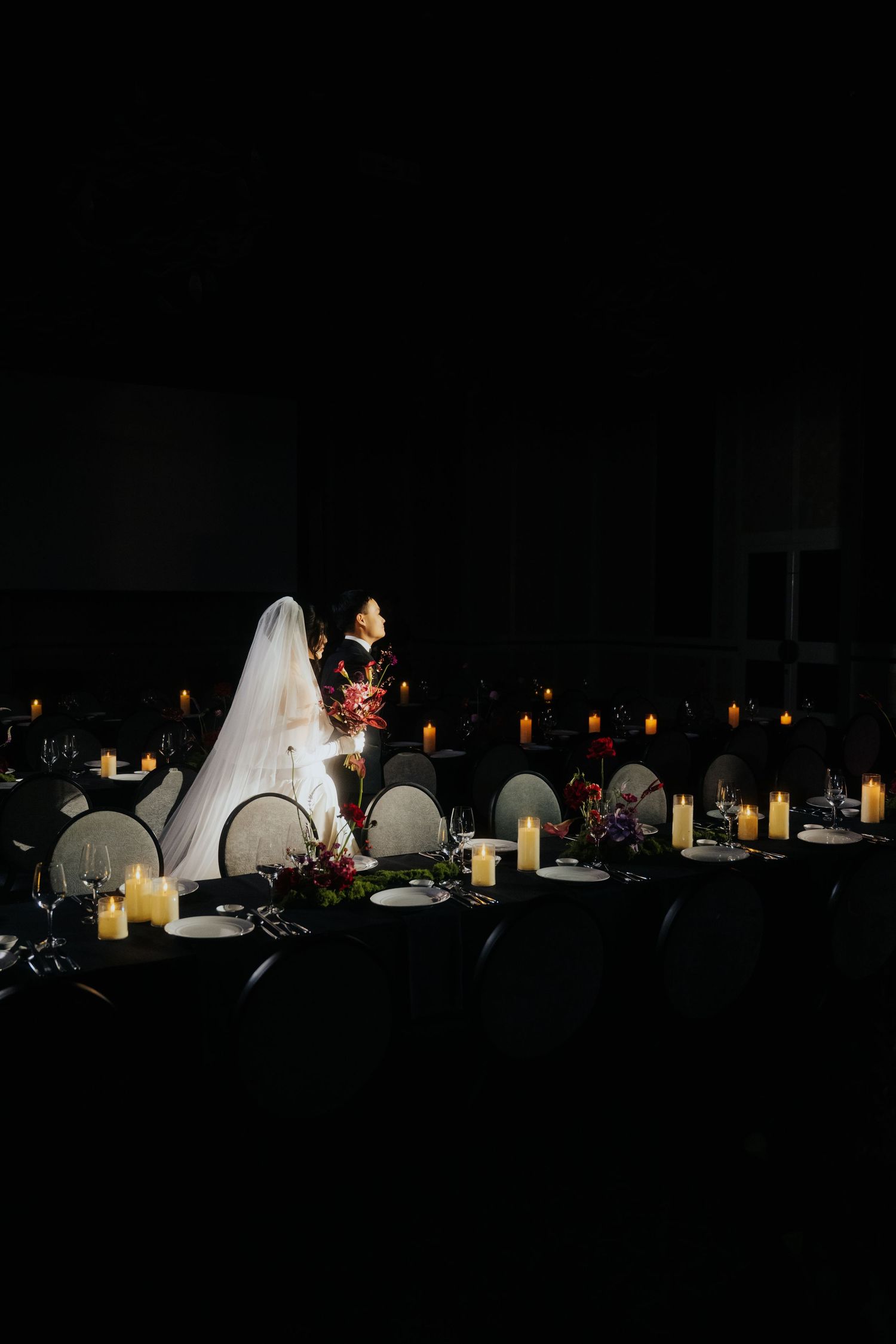 Bride and groom illuminated by candlelight in a dramatic dark reception hall with elegant black table settings.