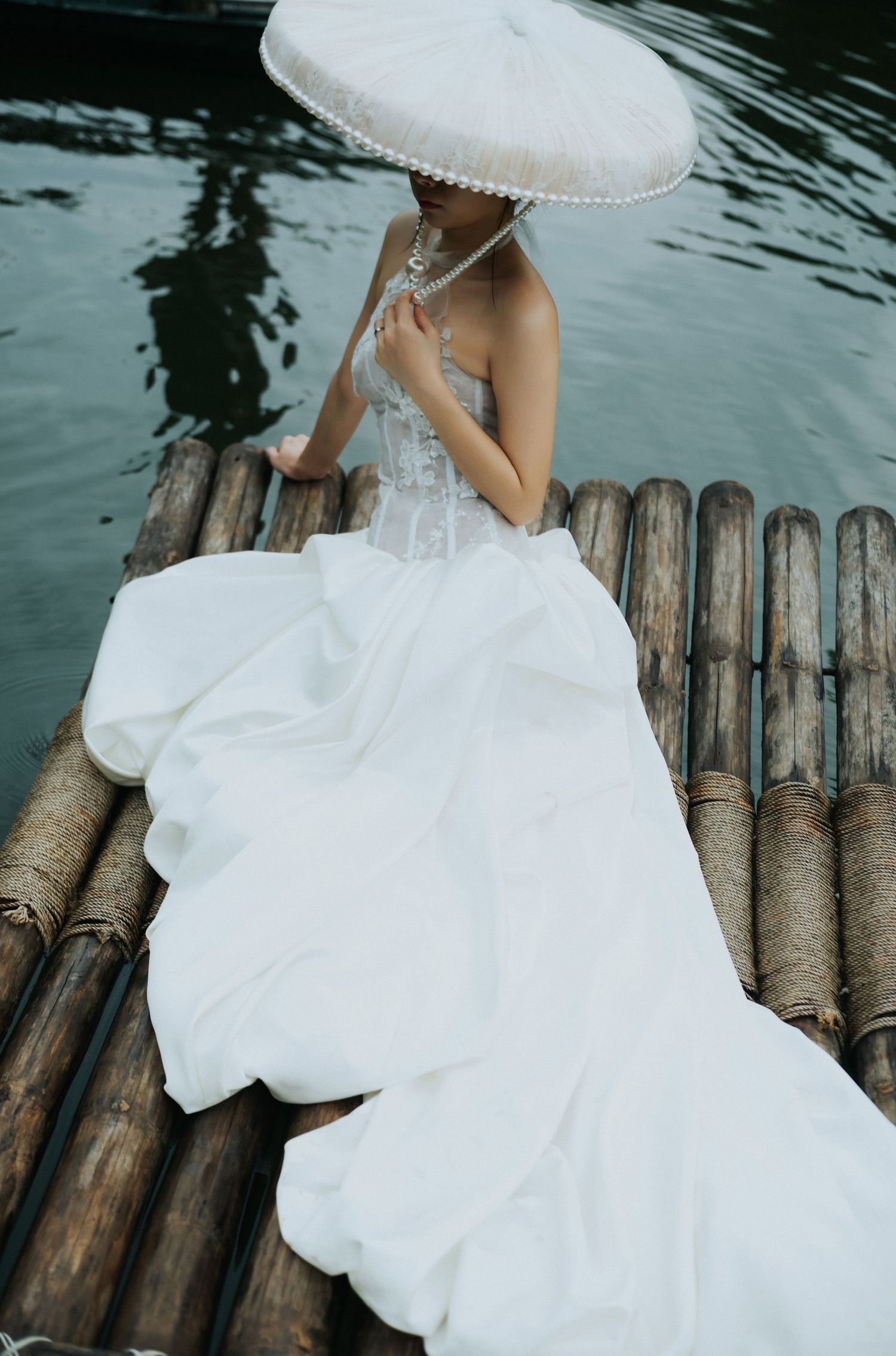 Bride in white wedding gown and parasol sitting on bamboo raft over calm water.