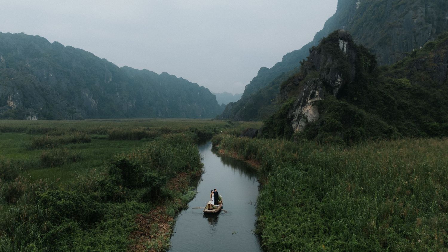 Aerial view of a small boat navigating a narrow river through lush green fields with dramatic karst mountains in Vietnam.