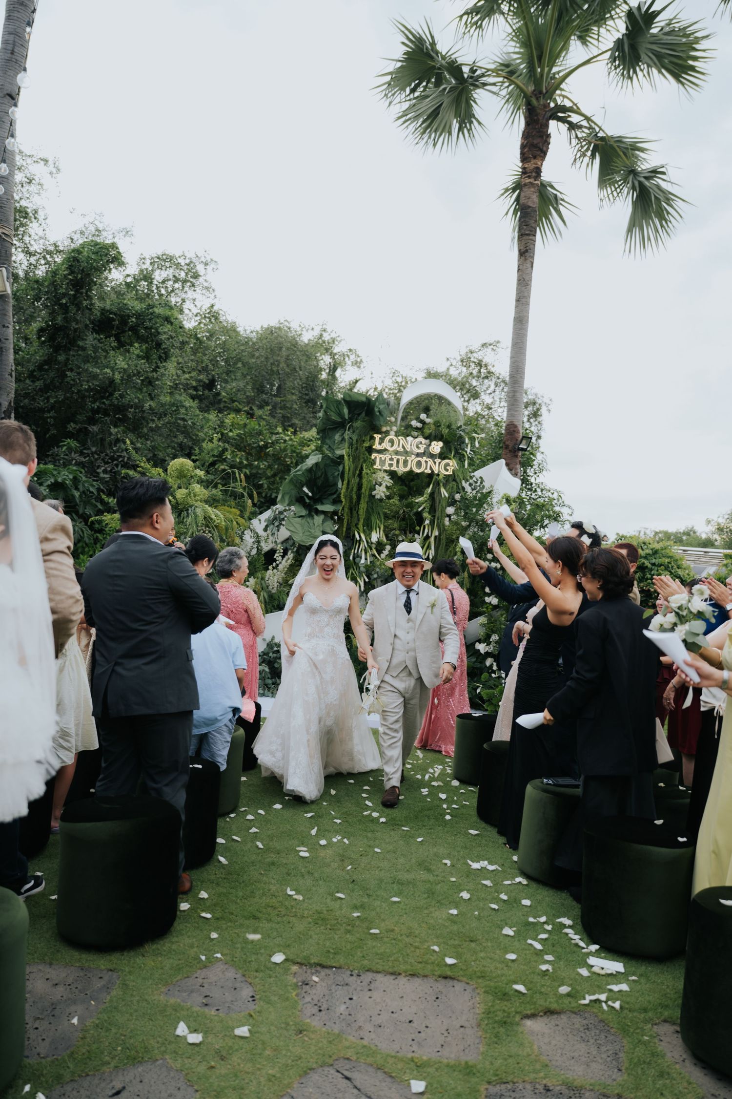 Bride and groom walk down outdoor aisle as guests celebrate with flower petals at a tropical garden wedding ceremony.