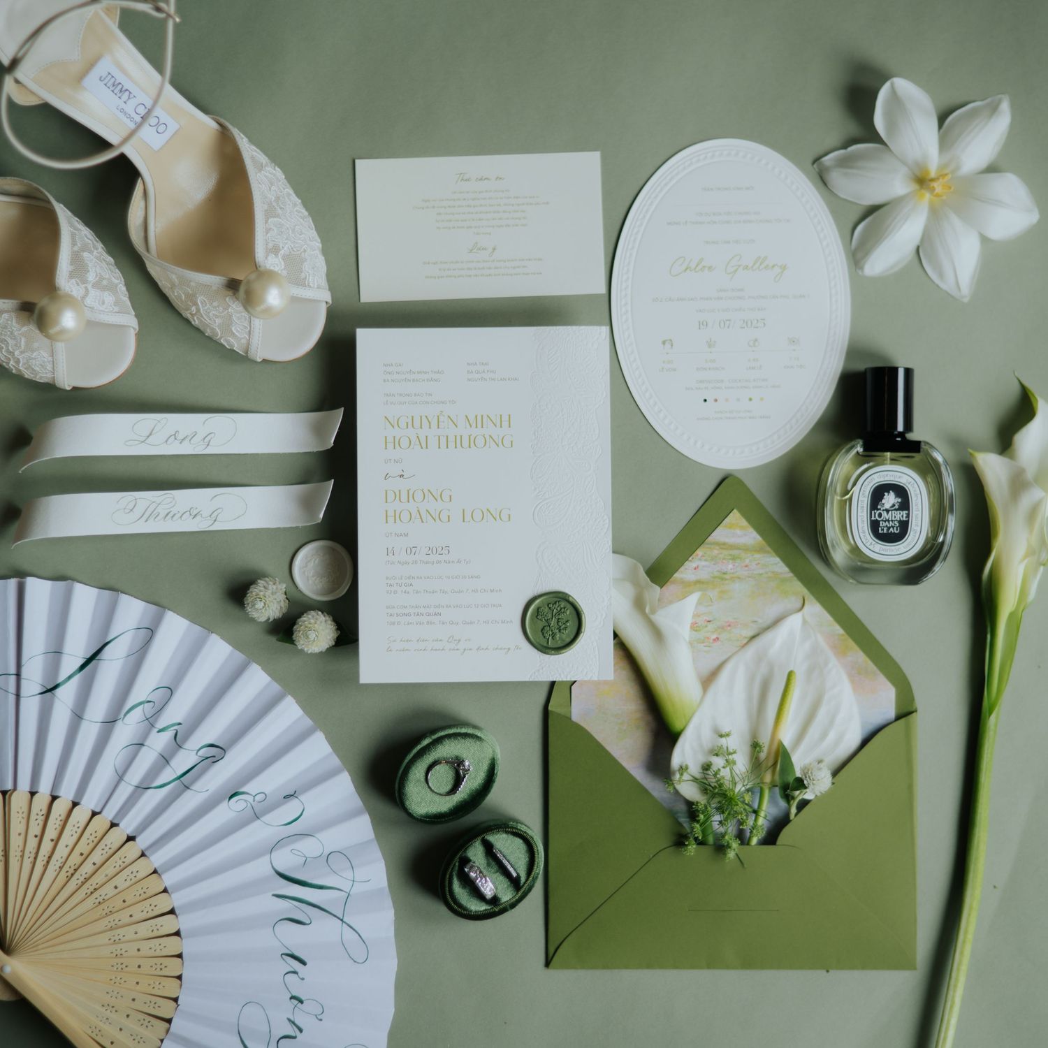 Elegant wedding stationery flatlay with green envelope, wax seal, fan, perfume, floral accents on sage green background.