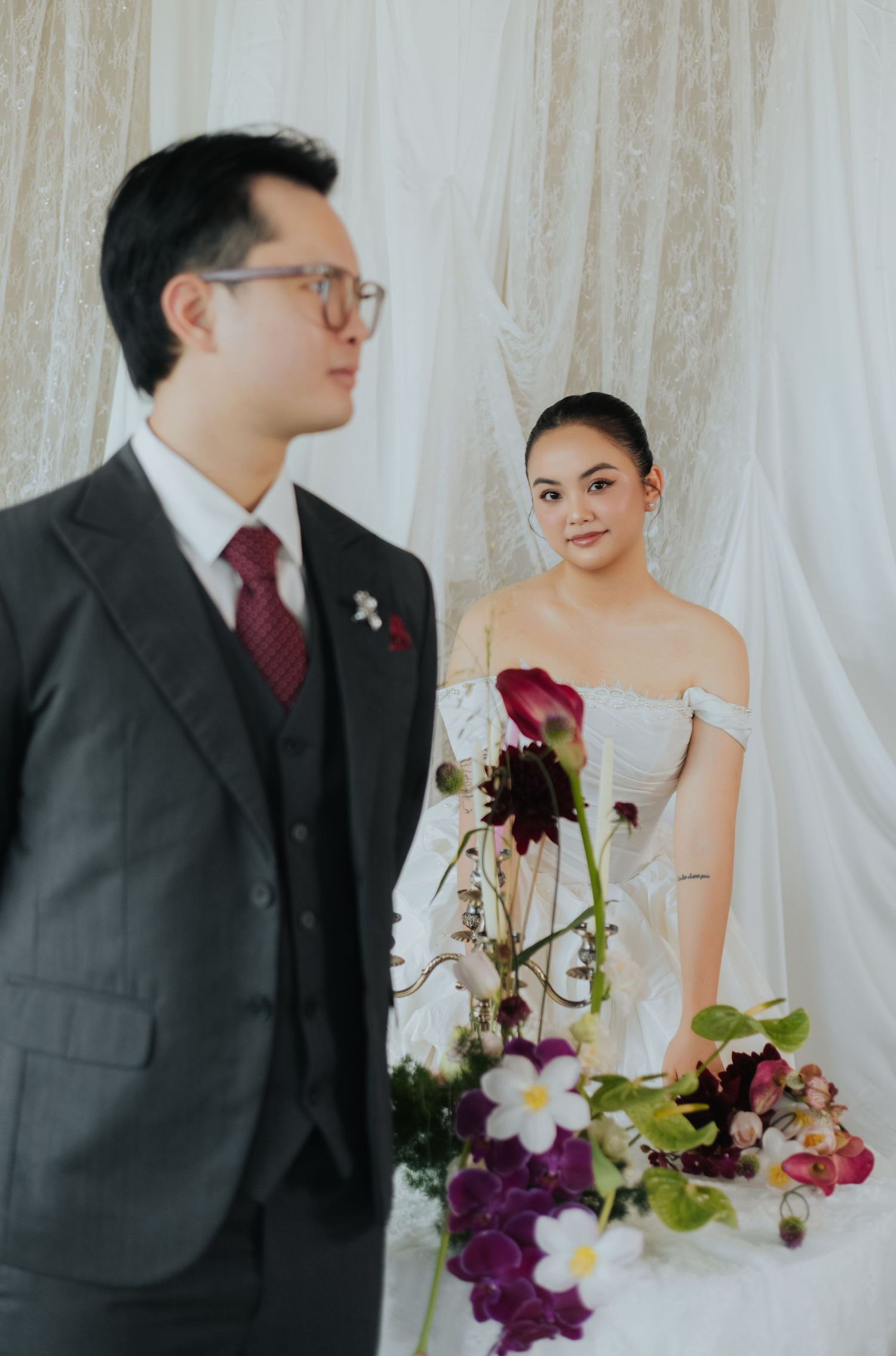 A bride in an off-shoulder white gown holds purple florals beside a groom in a gray suit with a red tie.