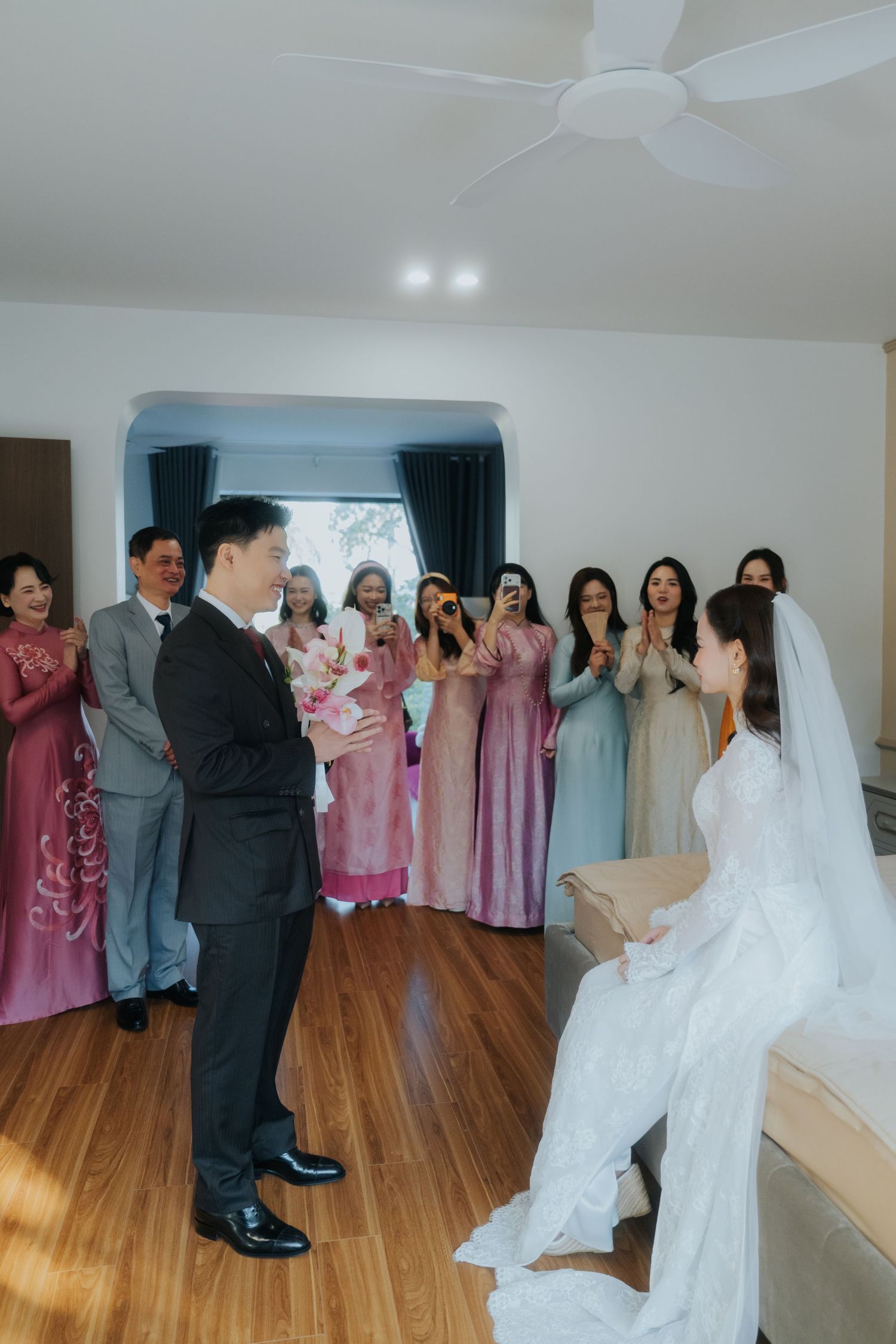 Groom presents bouquet to bride in white gown, surrounded by bridesmaids in pink ao dai at Vietnamese wedding.