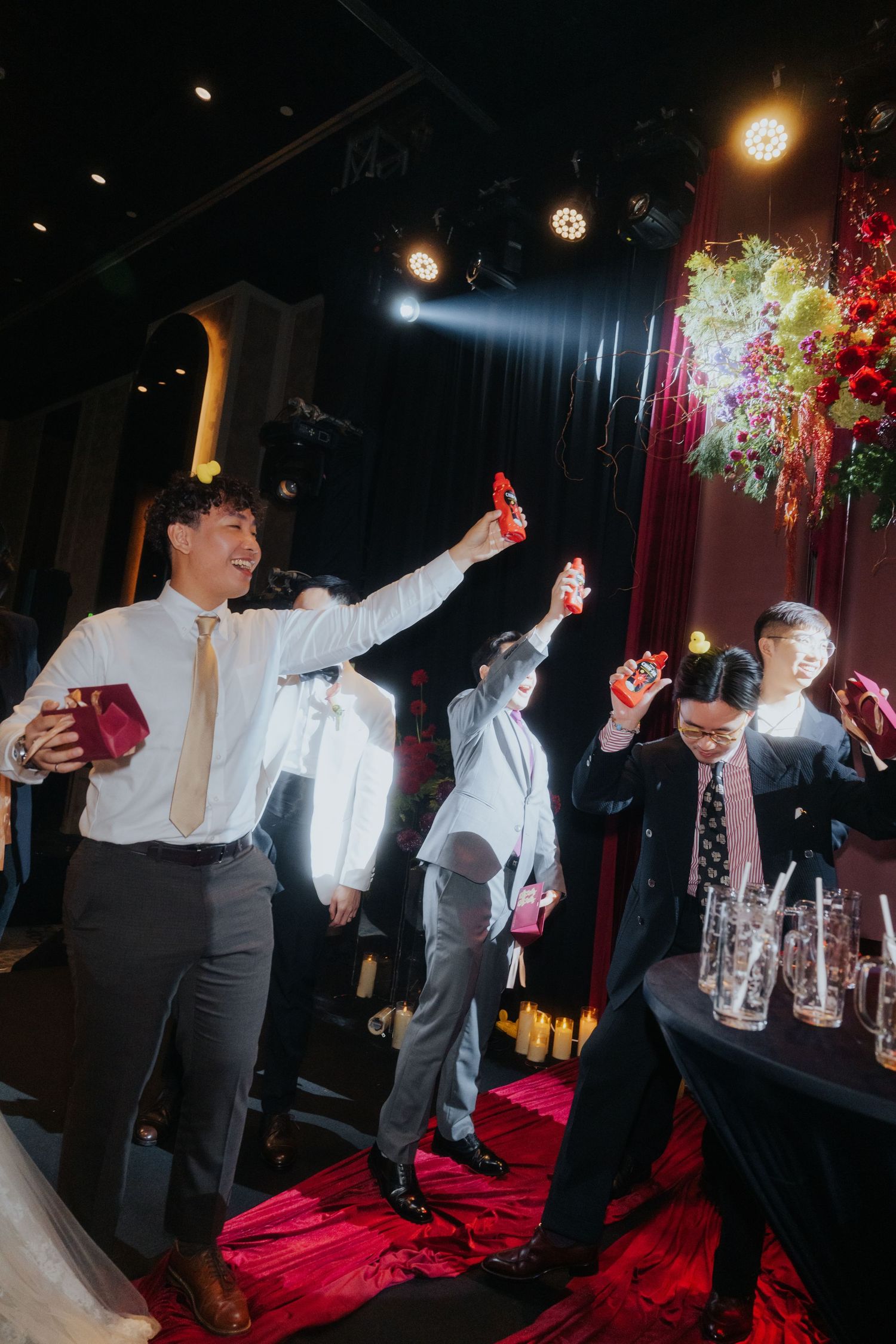 Guests in formal attire raise red gift boxes in celebration at an elegant indoor wedding reception event.