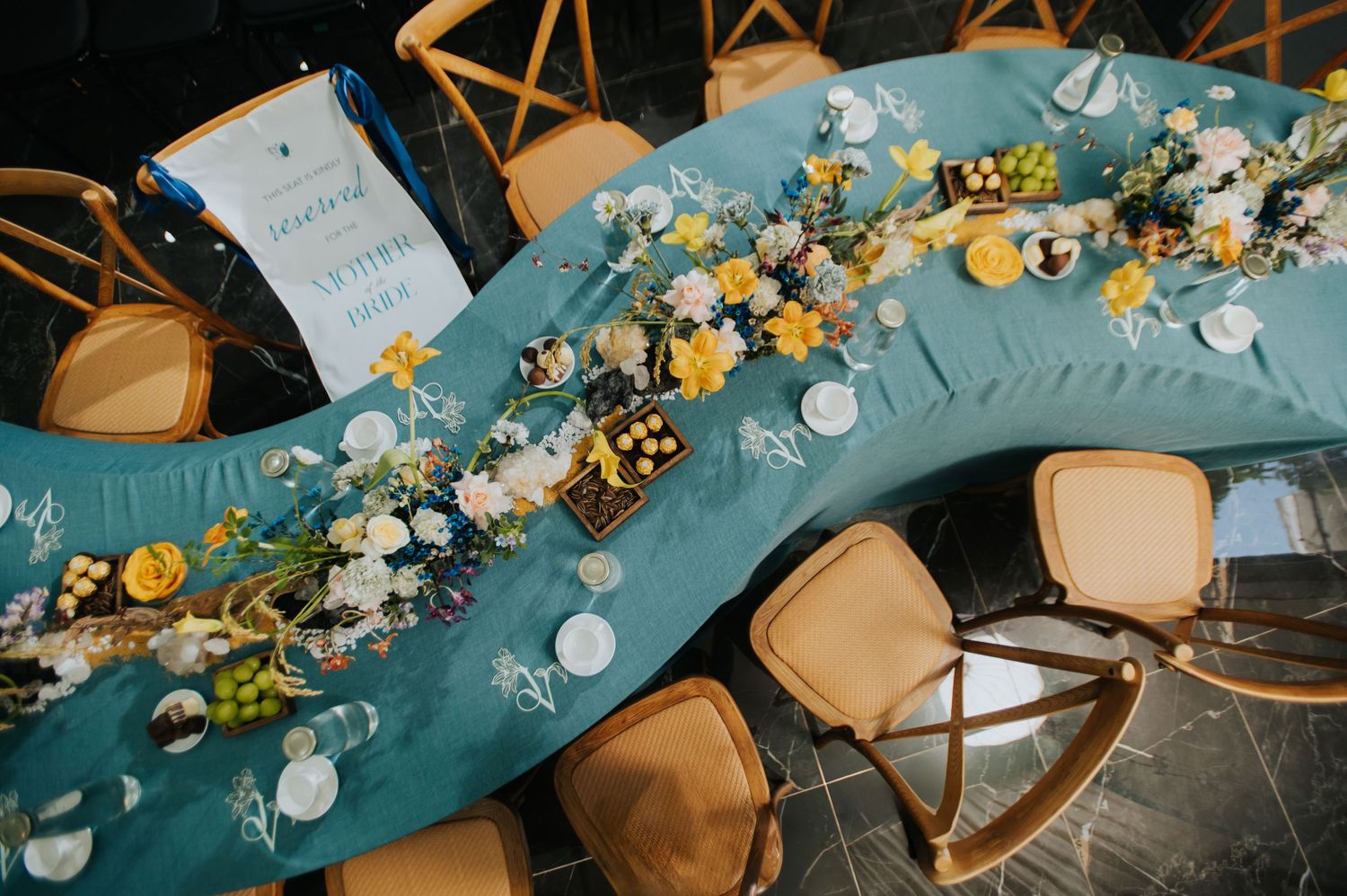 Aerial view of a curved banquet table with teal linen adorned with yellow and white floral arrangements and wooden chairs.