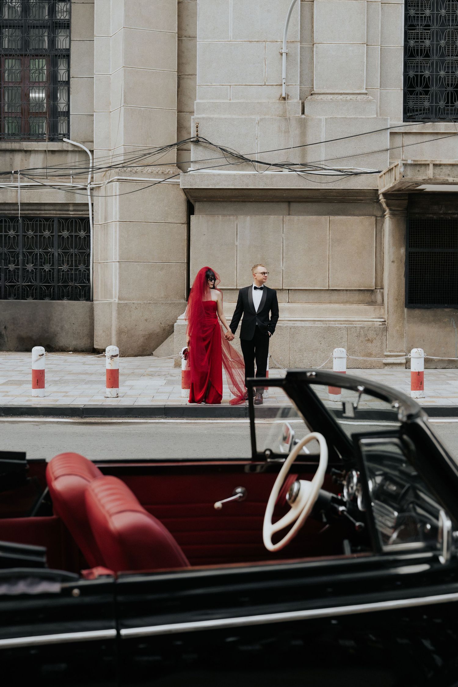 Elegant couple in formal attire, woman in red gown and man in tuxedo, standing near a vintage black convertible car.