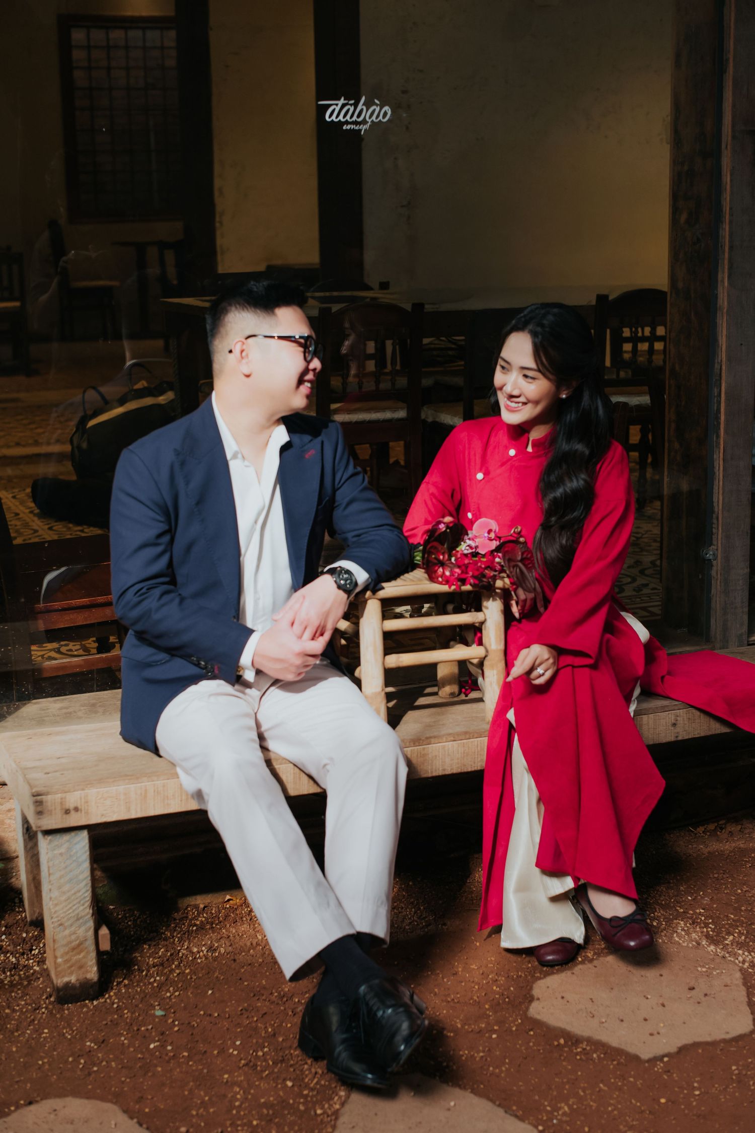 A couple sits on a wooden bench, man in navy blazer and woman in red ao dai holding flowers, sharing a joyful moment.