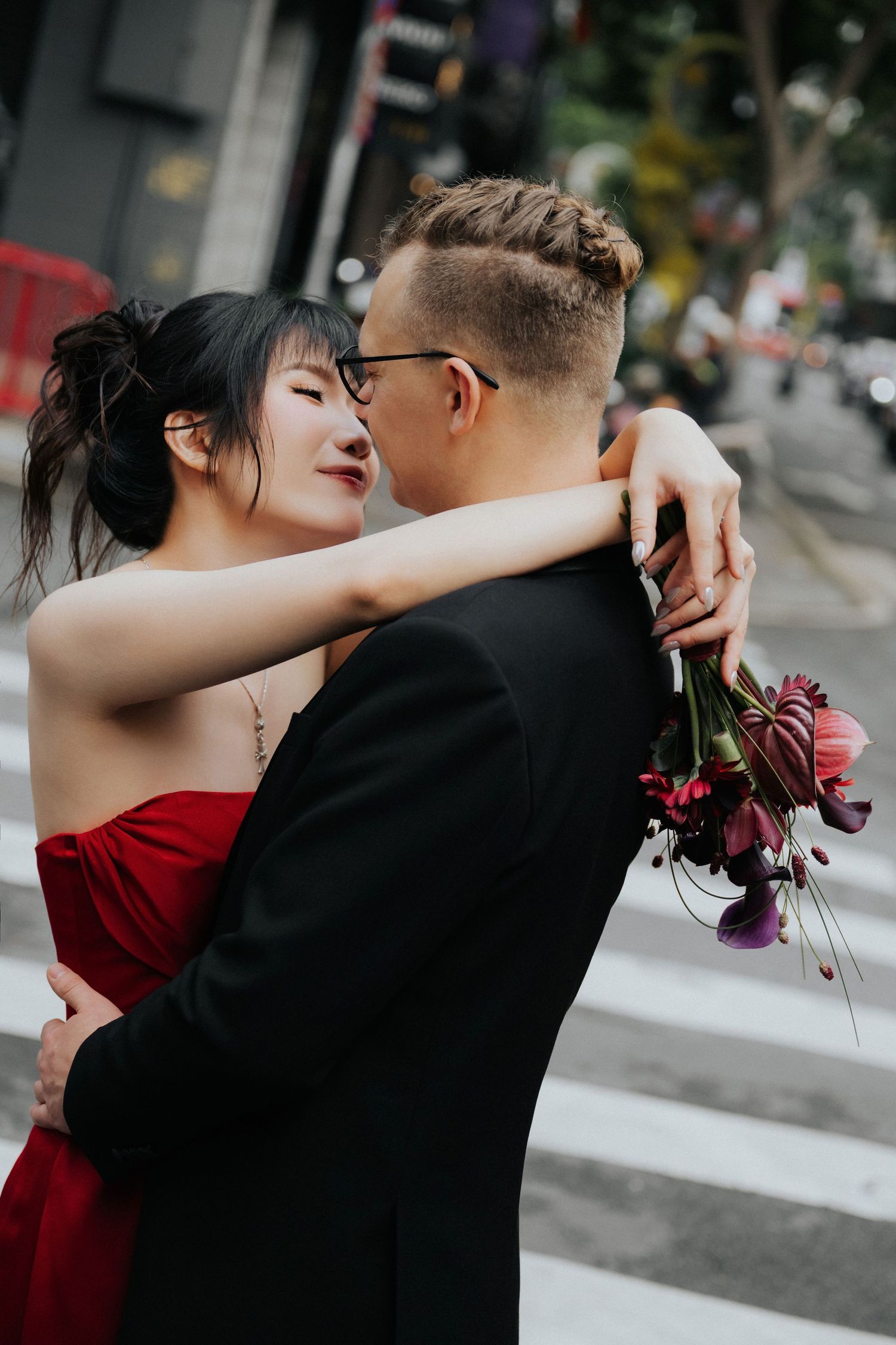 Couple embracing on a city crosswalk, woman in red strapless gown holding dark floral bouquet, man in black suit.
