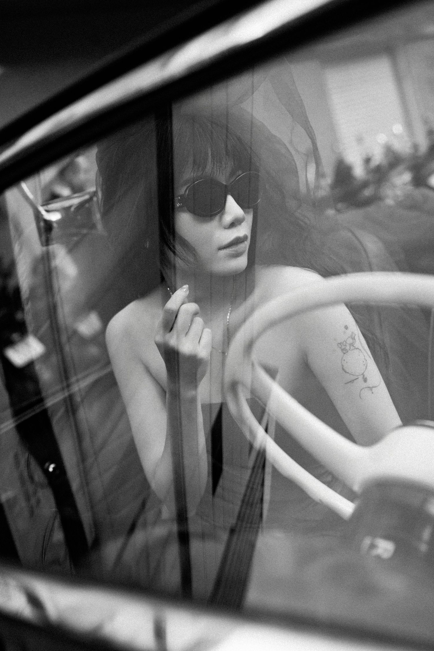 Black and white photo of a woman with sunglasses and tattoos sitting in a vintage car, viewed through the window.