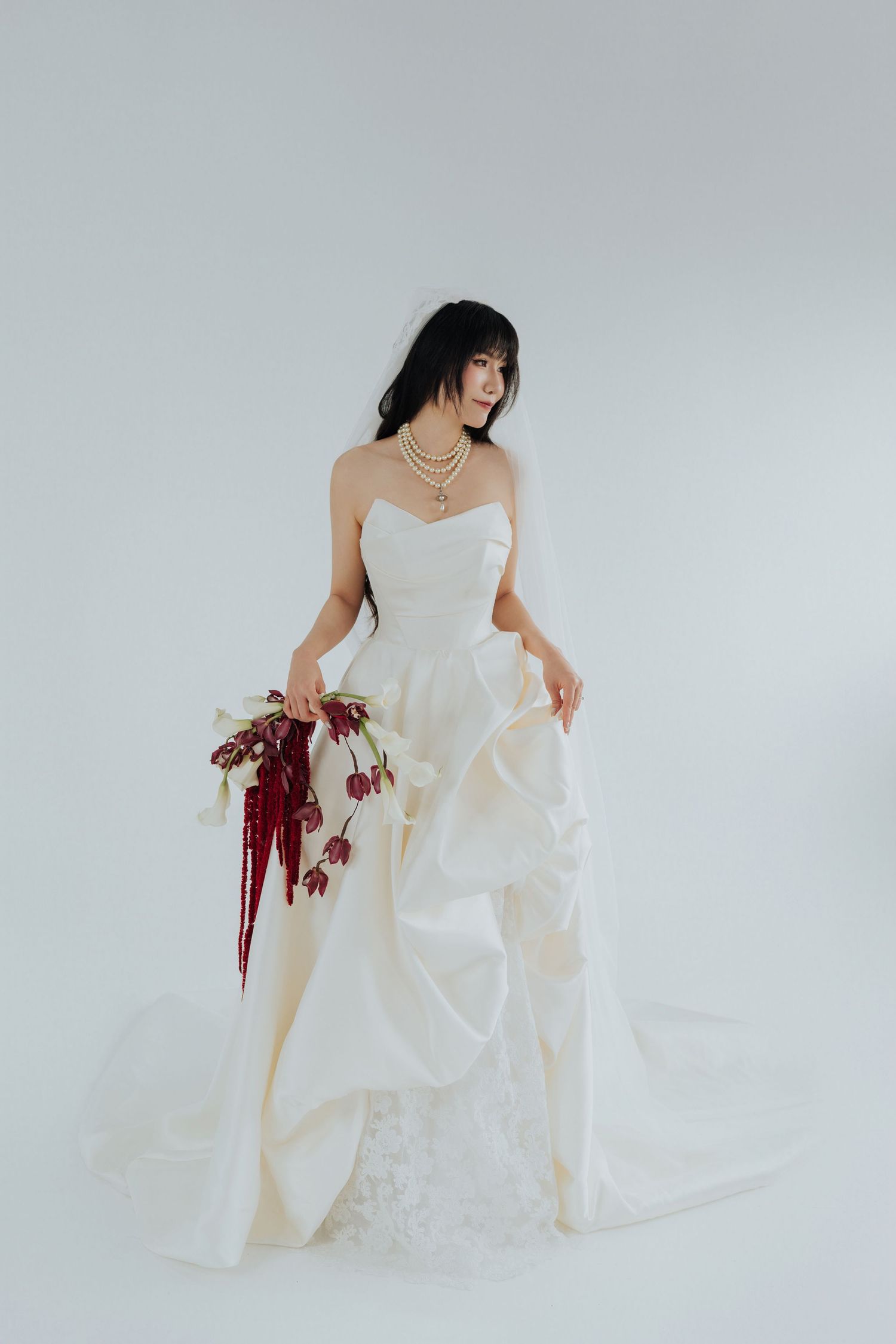 Bride in strapless white ballgown with ruffles holds red floral bouquet, wearing pearl necklace on white background.